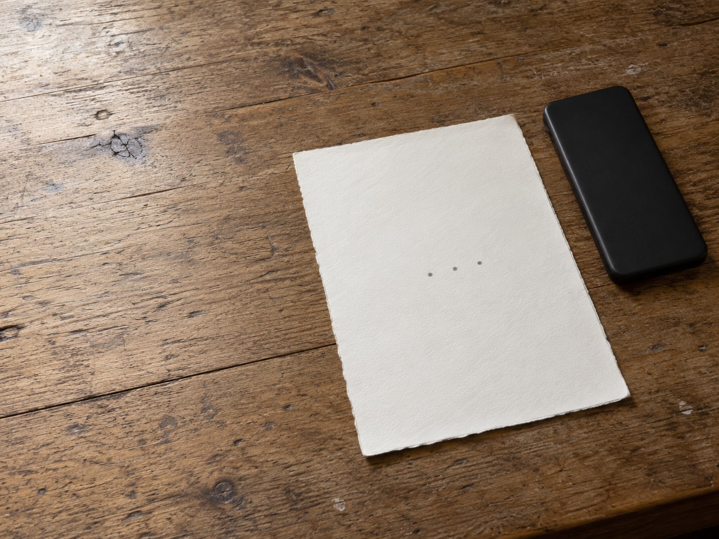 A blank sheet of paper with three small pencil dots, a closed phone lying face down beside it on a wooden counter.