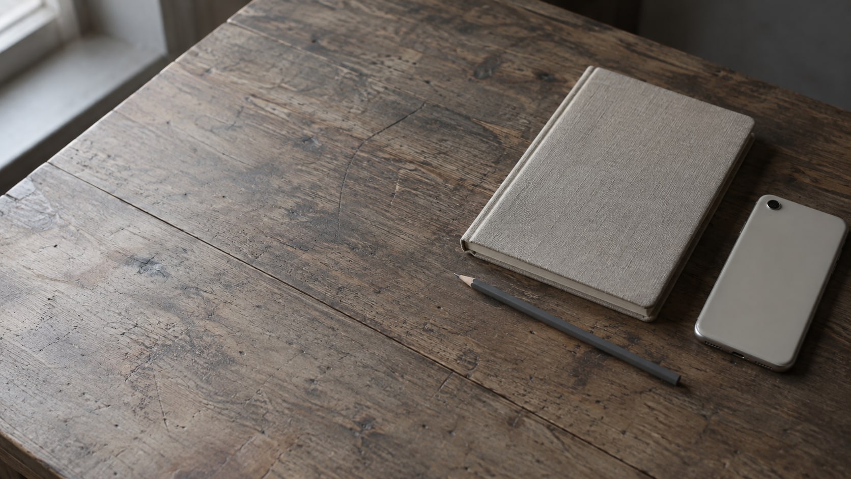 A closed notebook, a pencil and a face-down phone arranged on a worn wooden desk.