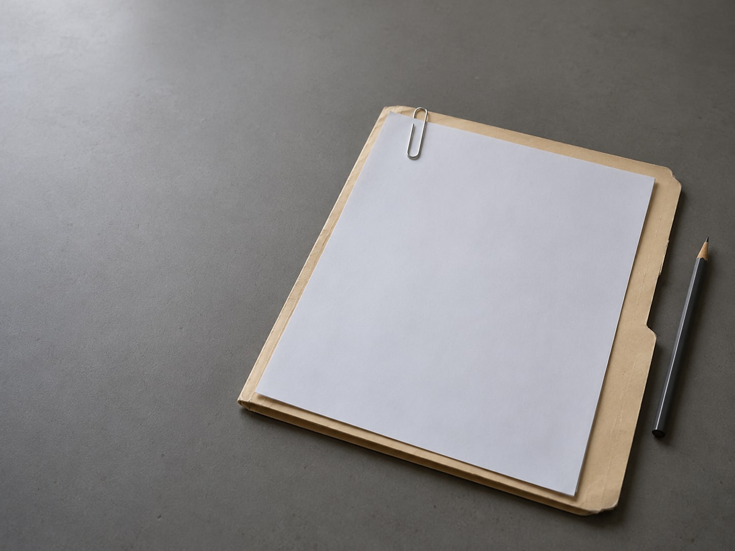 A closed manila folder with a blank sheet held by a paper clip, a pencil beside it, on a plain gray desk.
