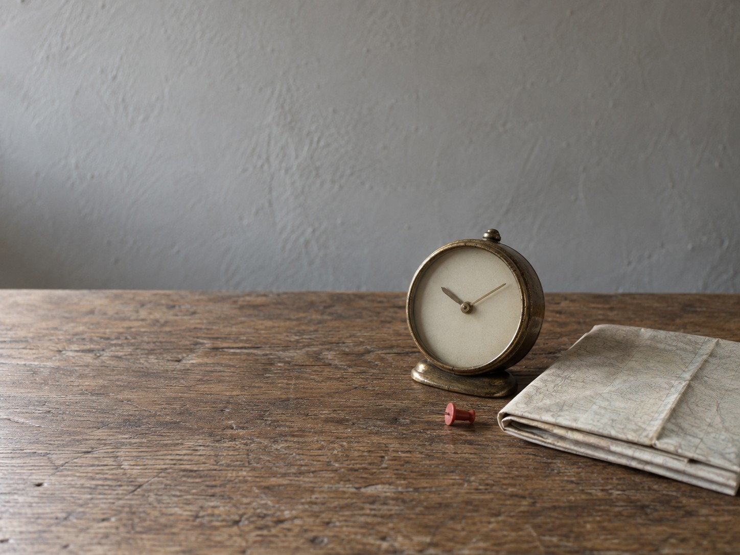 A brass desk clock beside a closed paper map and a single pushpin resting on a bare oak table.