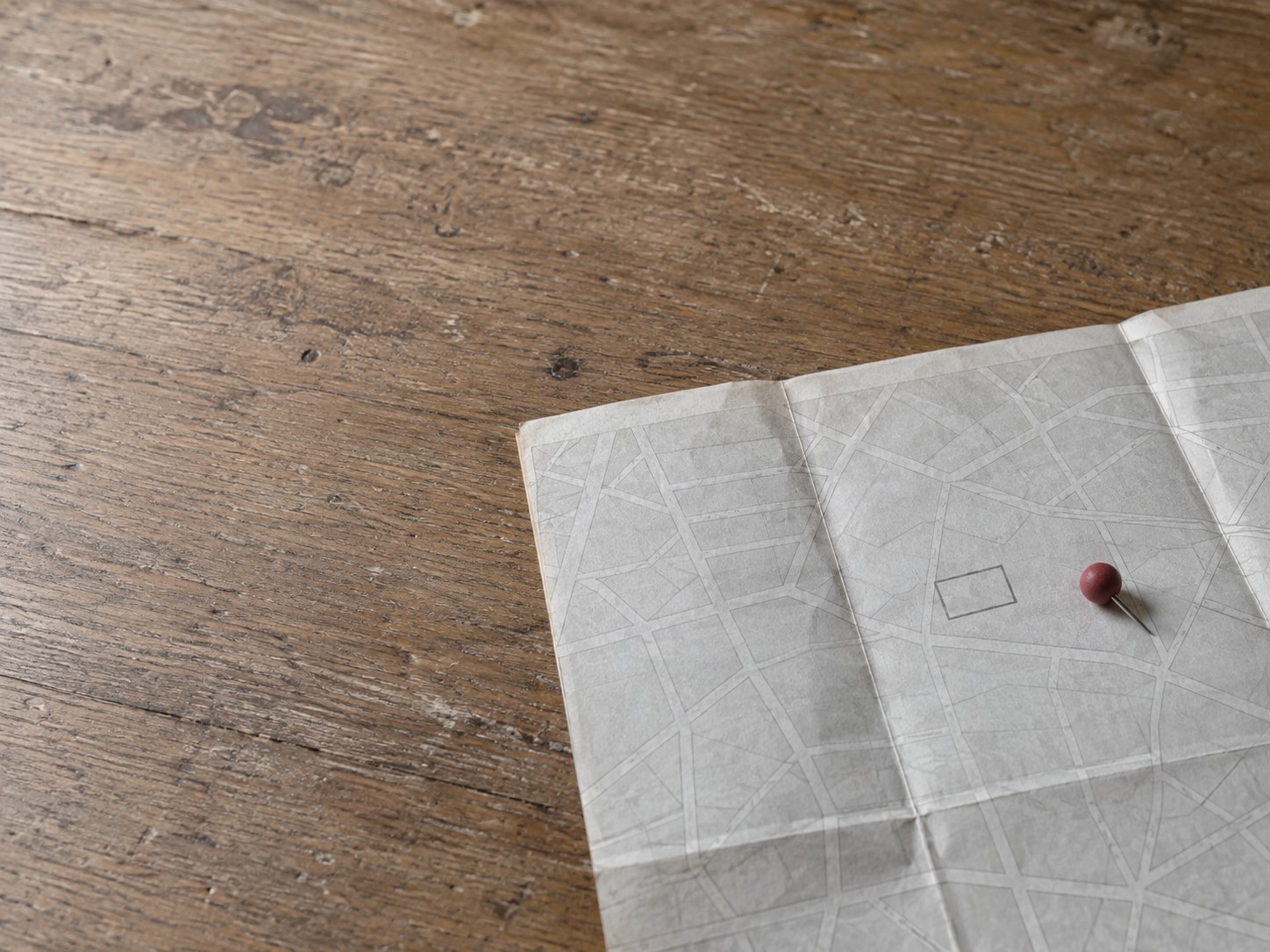 A folded paper street map on a wooden counter with a single red pushpin set slightly off to one side of a drawn building outline.