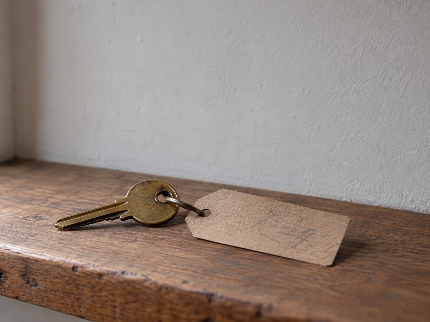 A worn brass key beside a small paper tag with pencil scribbles on a wooden shelf.