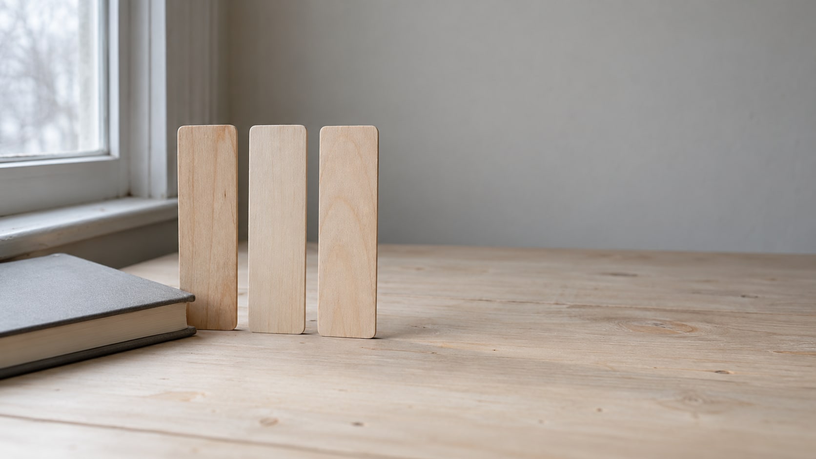 A row of three blank wooden bookmarks standing upright on a pale desk beside a closed hardcover notebook.