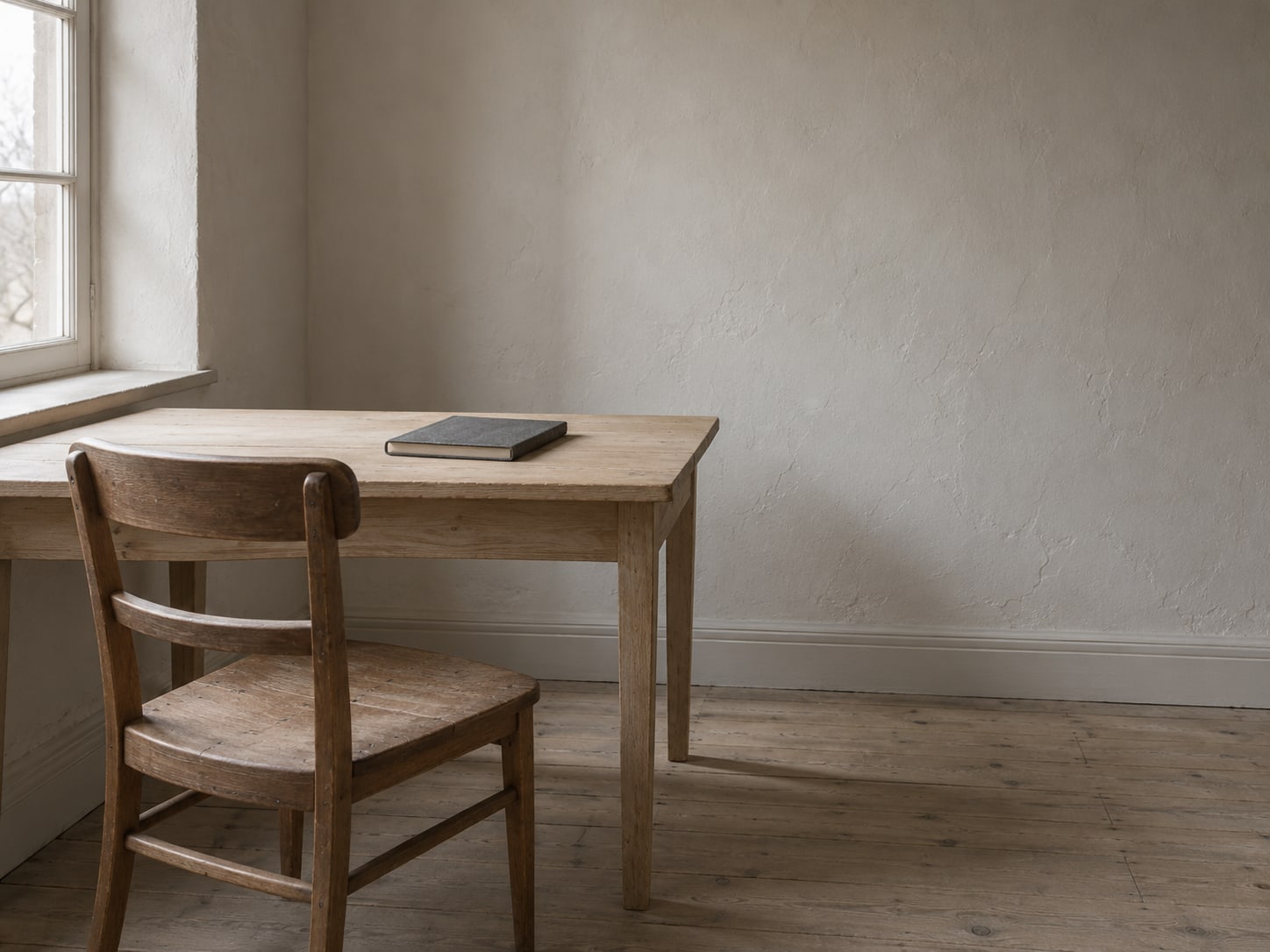 A single wooden chair facing an empty desk with a closed notebook on it, in a quiet room.