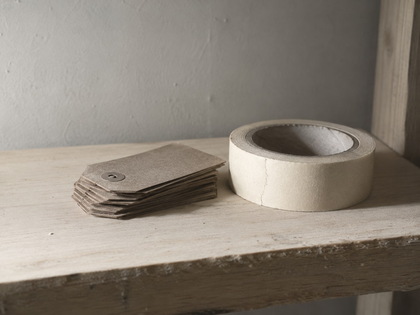 A small stack of plain cardboard shipping tags beside a roll of masking tape on a pale wooden shelf.