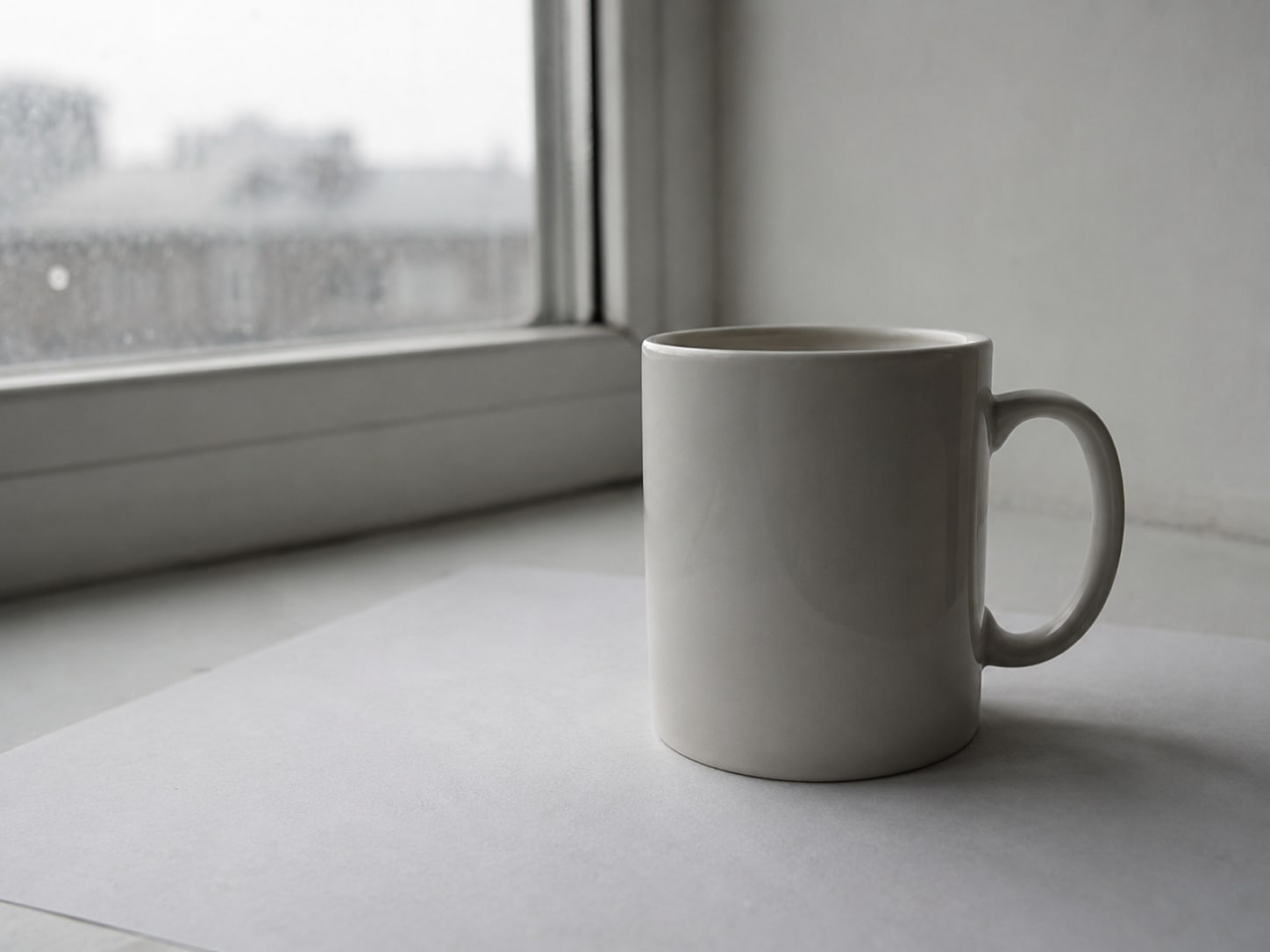A plain white ceramic mug on a sheet of white paper beside a window, with soft daylight from one side.