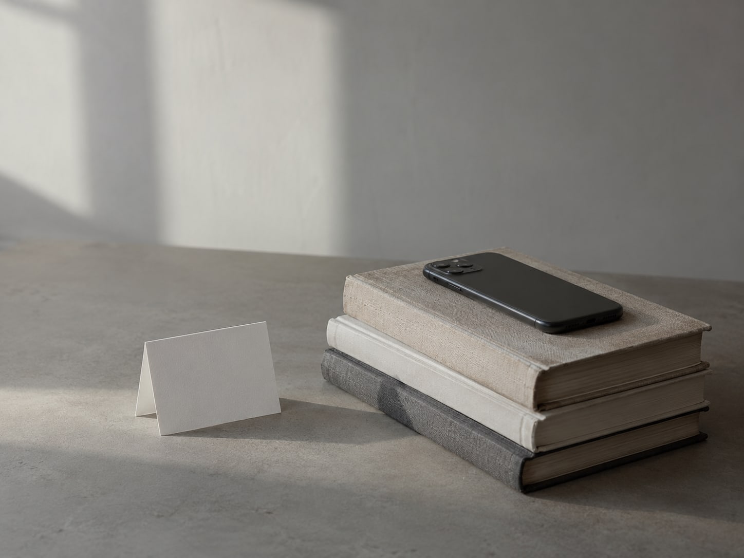 A phone lying face down on a stack of three books beside a small folding white card, on a bare table.