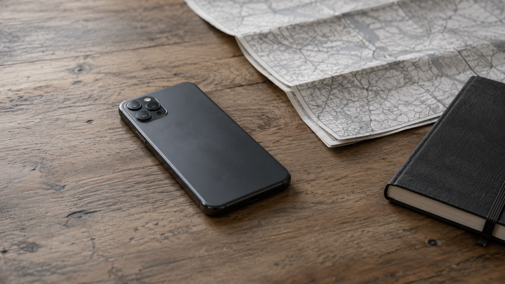 A smartphone lying face down on a wooden desk beside a folded paper map and a closed notebook.