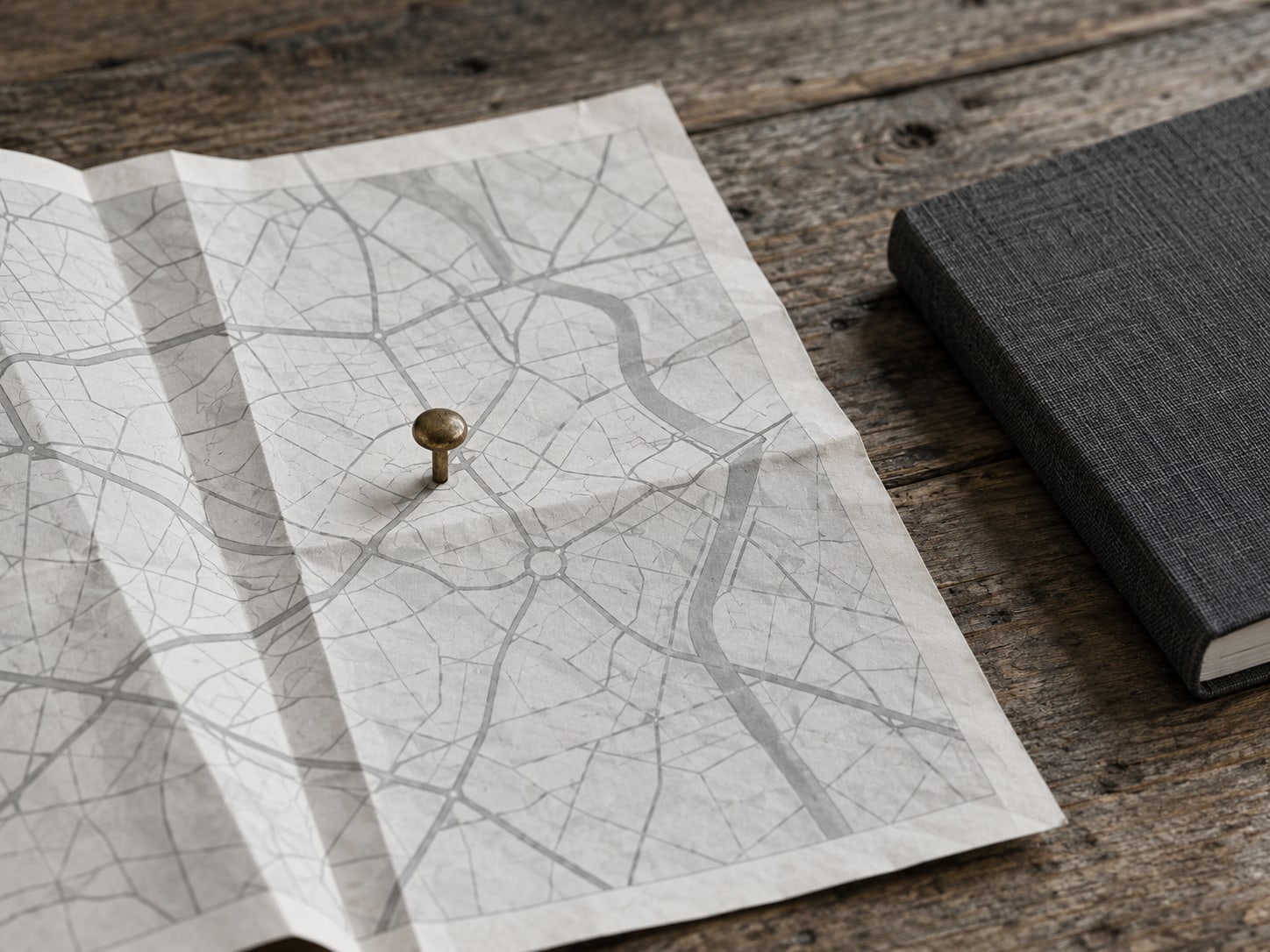 A folded paper street map with one brass pushpin on a wooden table beside a closed notebook.