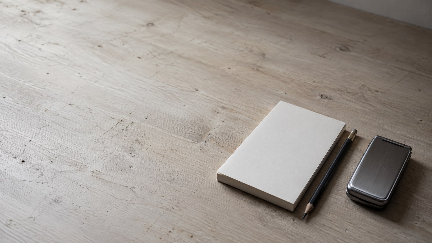 A closed flip phone, a blank notepad and a pencil arranged on a pale wooden counter.