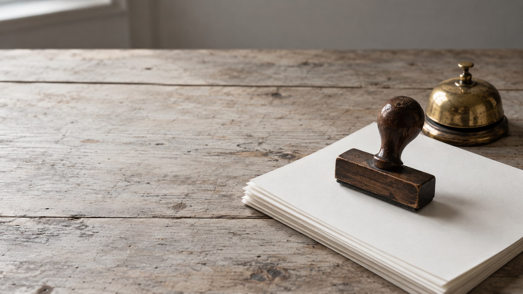 A single rubber stamp resting on a stack of blank sheets beside a small brass bell on a plain table.
