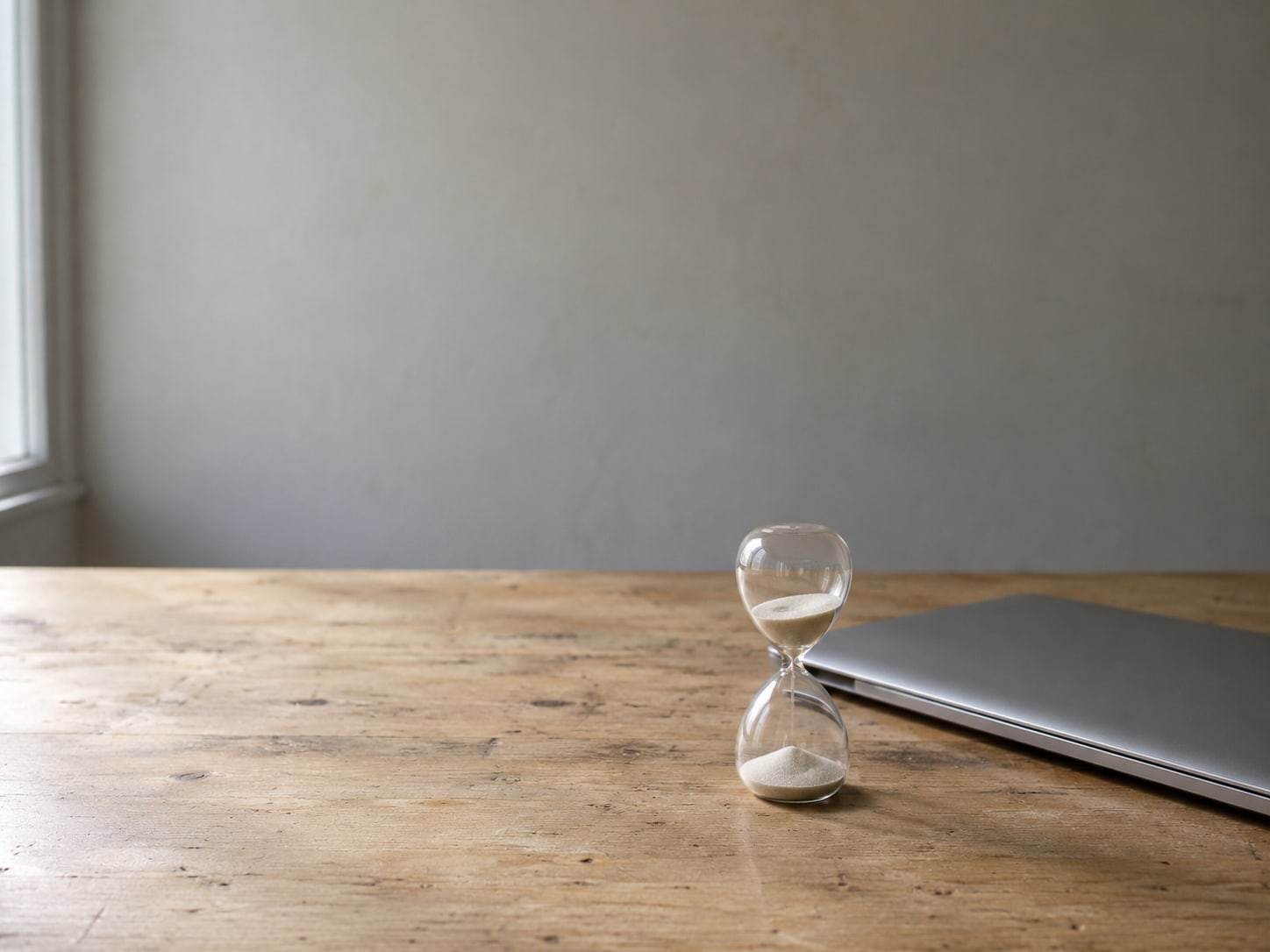 A small hourglass beside a closed laptop on a plain wooden desk.