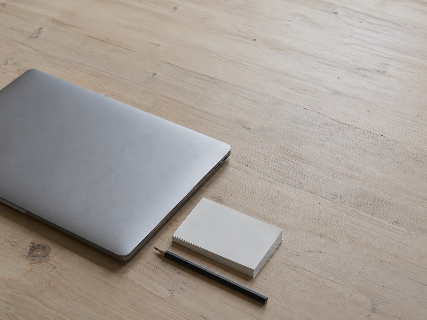 A closed laptop beside a small stack of blank cards and a pencil on a light wooden desk.