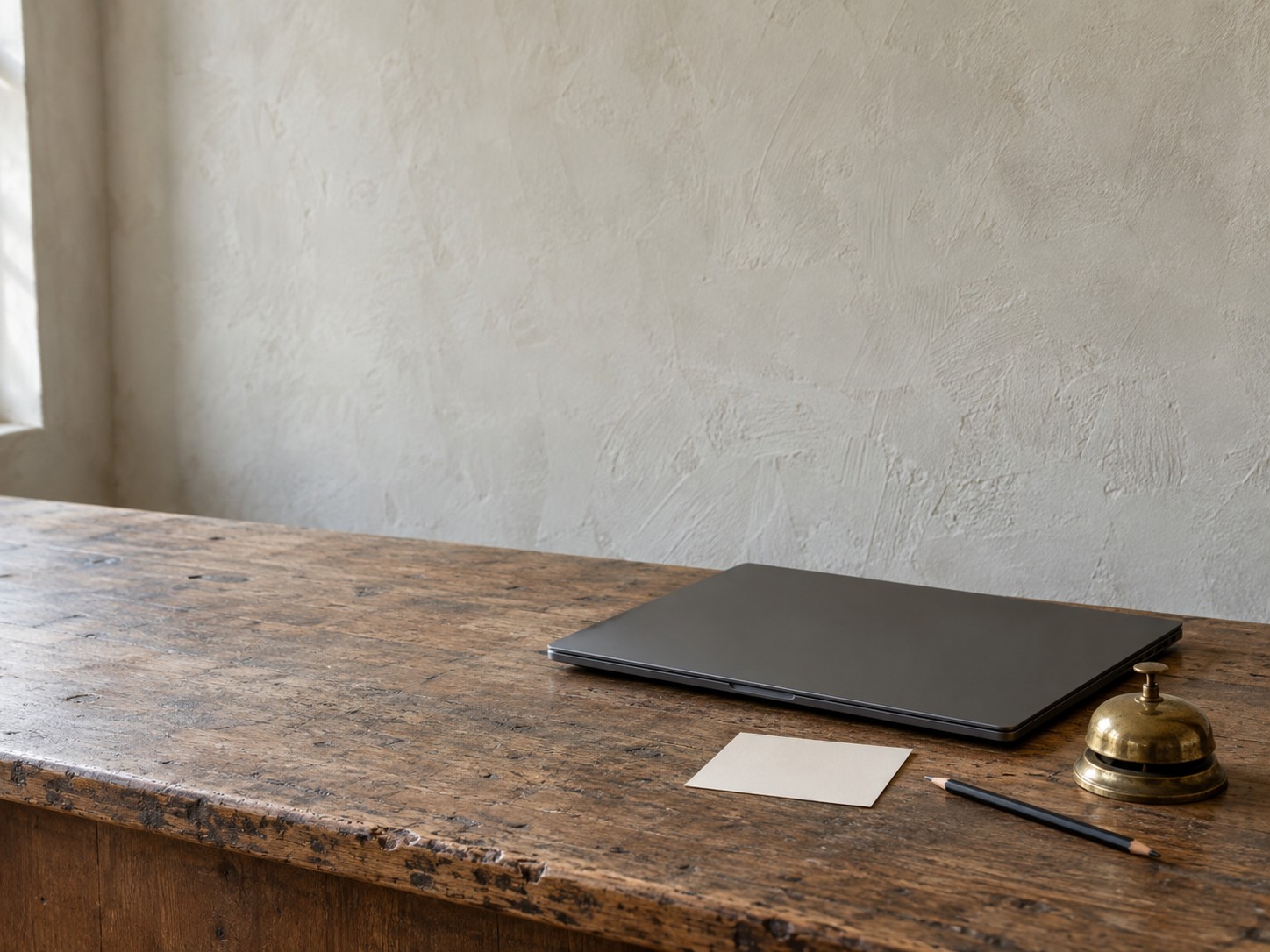 A closed laptop, a blank index card and a pencil beside a small brass desk bell on a shop counter.