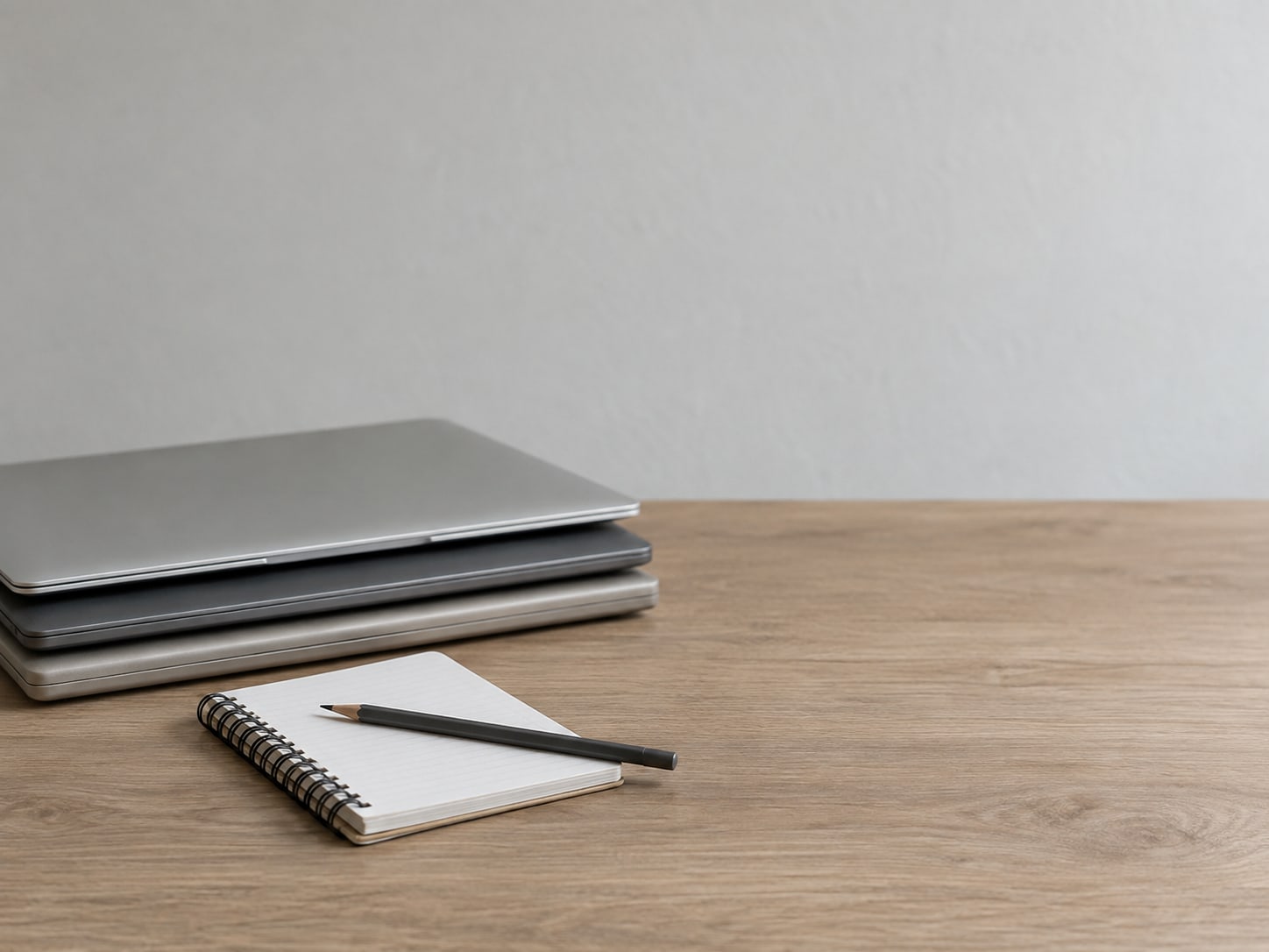 Three closed laptops stacked neatly on a desk beside a plain spiral notebook with a pencil resting on it.