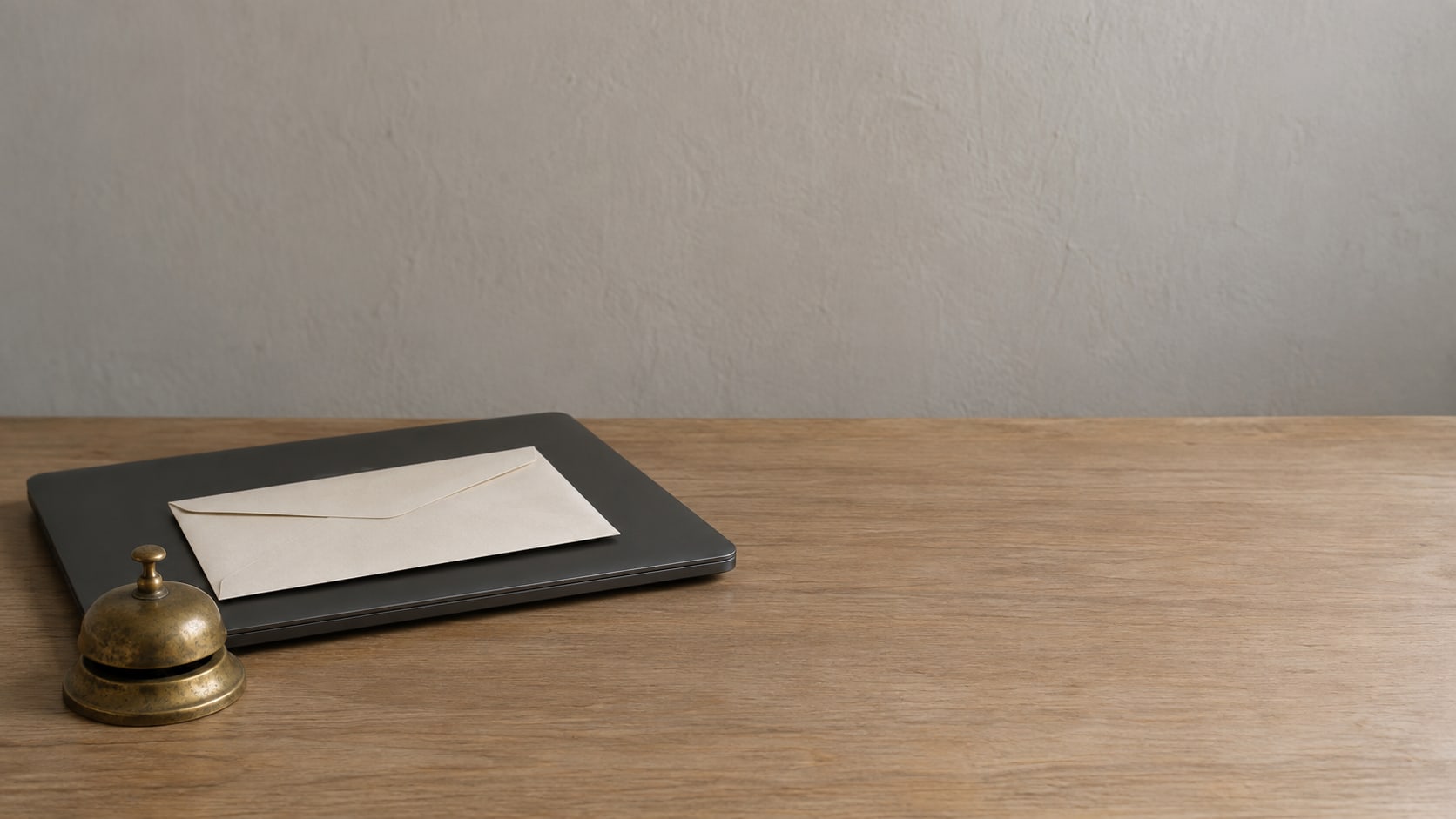A sealed envelope resting on a closed laptop on a wooden desk beside a small brass desk bell.