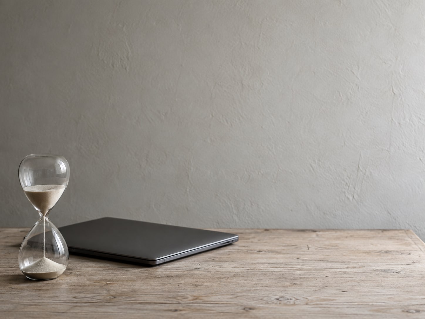 An hourglass with sand running through it, standing beside a closed laptop on a pale desk.