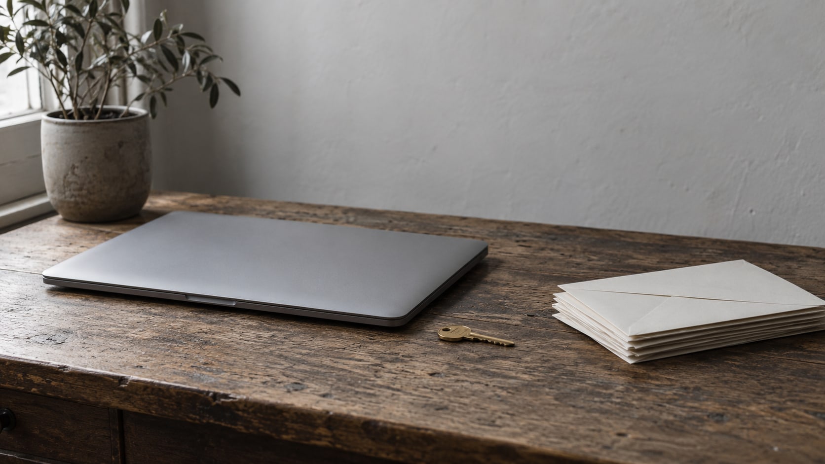 A closed laptop beside a small brass key and a stack of unopened envelopes on a worn wooden desk.
