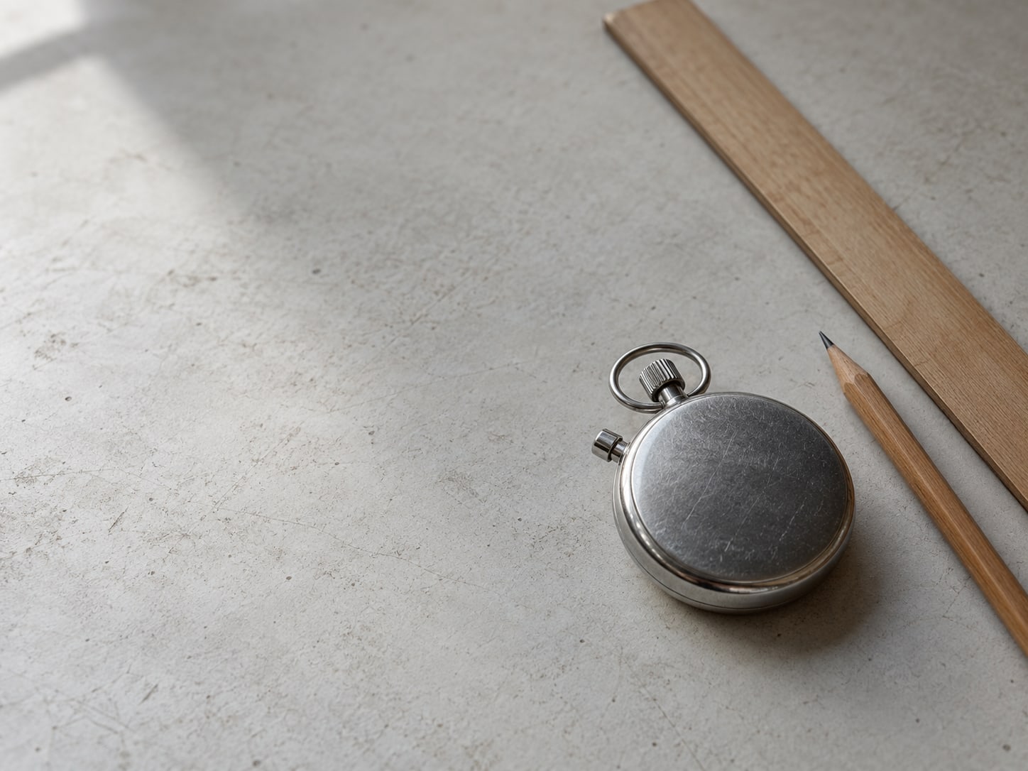 A stopwatch lying face down on a desk beside a plain wooden ruler and a pencil.