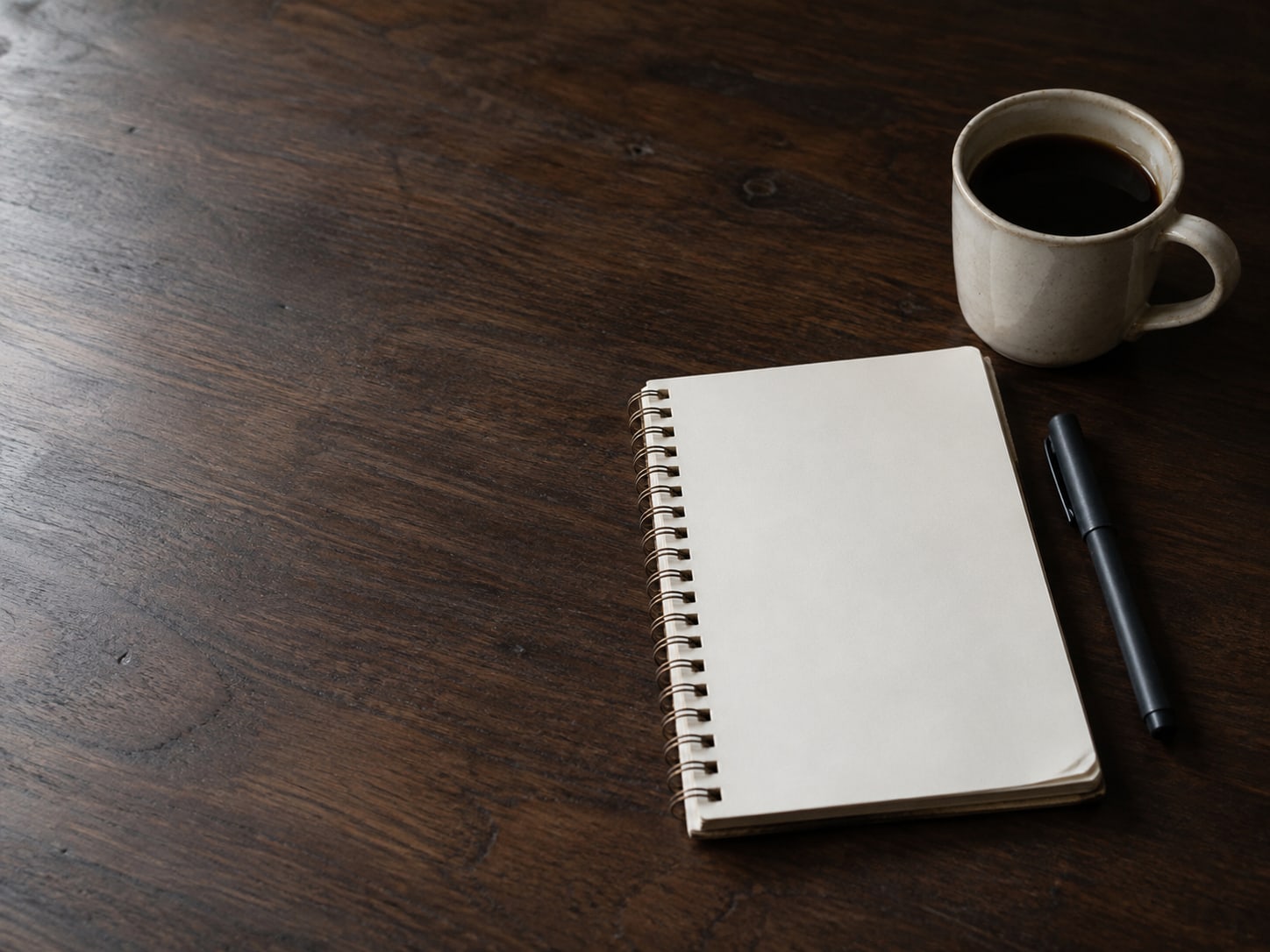 A spiral notebook open to a blank page next to a capped pen and a mug of coffee on a dark walnut table.