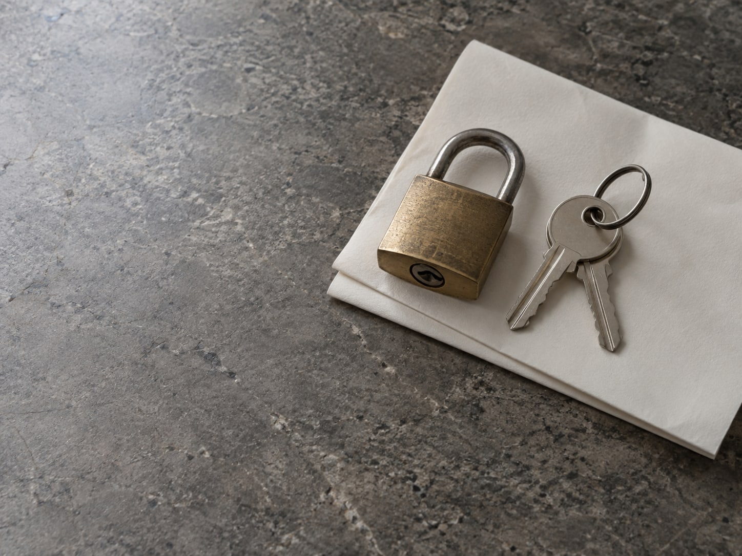 A small brass padlock resting on a folded sheet of plain paper beside a set of keys on a gray stone counter.
