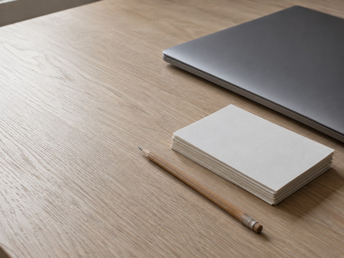 A closed laptop beside a short stack of blank index cards and a pencil on a pale oak desk.