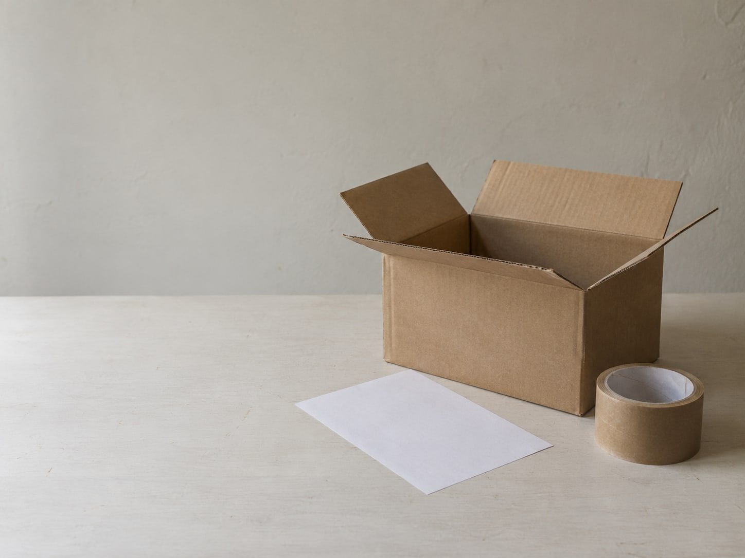 Una caja de cartón de envío con las solapas abiertas, un comprobante de devolución en blanco y un rollo de cinta adhesiva sobre una mesa limpia.