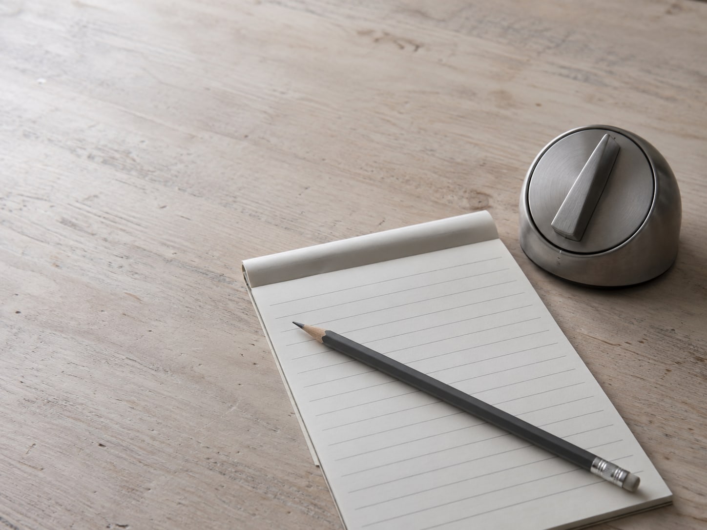 A pencil lying across a blank lined checklist pad beside a kitchen timer set on a wooden table.