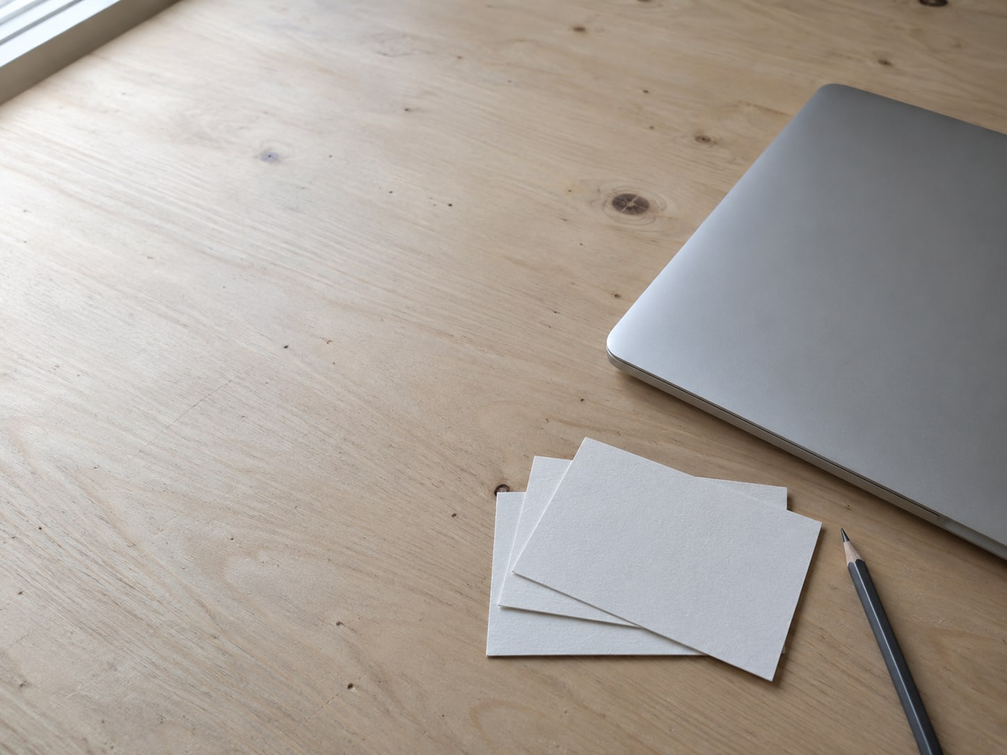 A pale wooden desk with a closed laptop and three blank index cards fanned out beside a pencil, ready for a page-by-page plan.