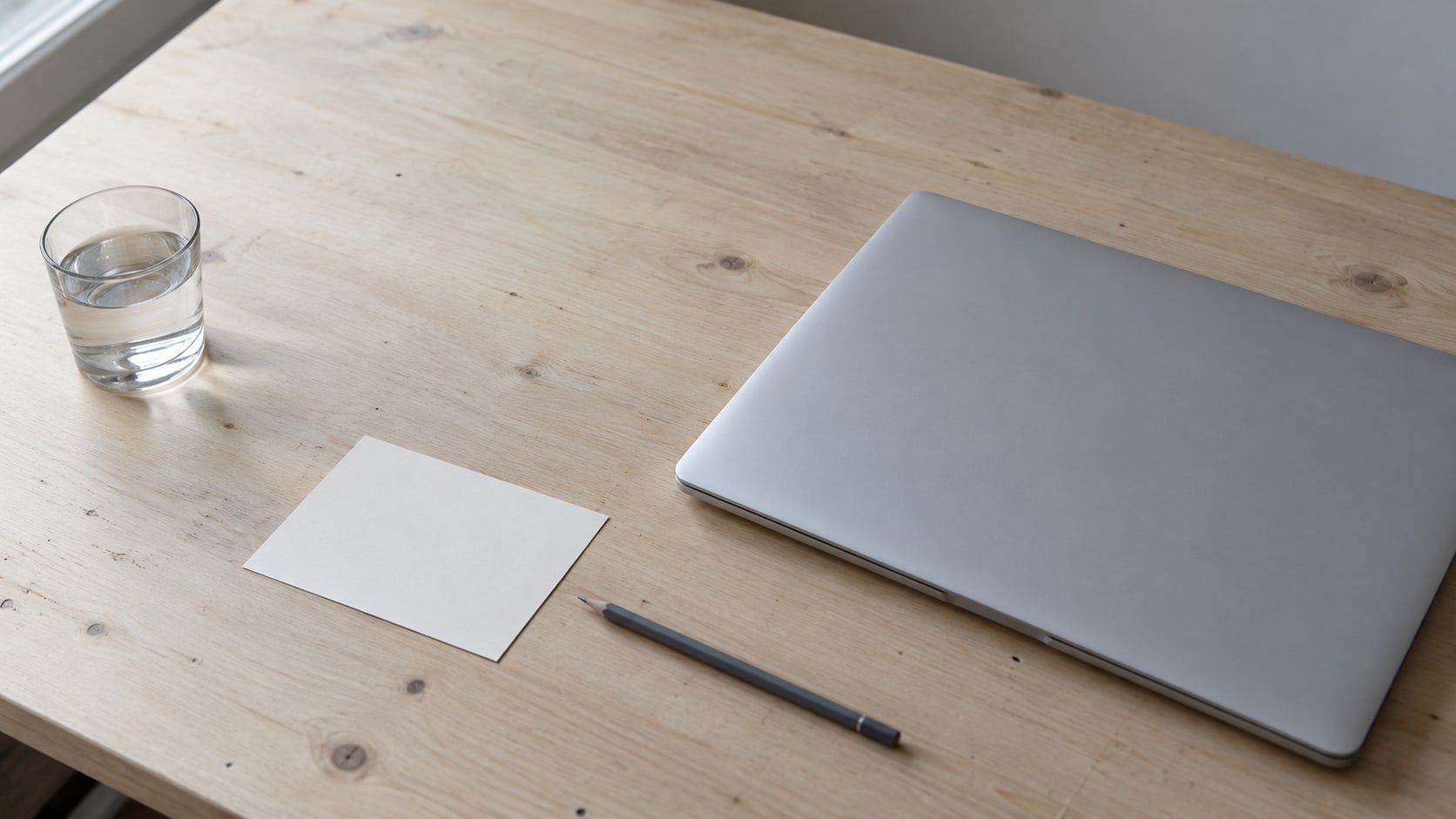 A pale wooden desk with a closed laptop and a blank index card beside a pencil and a small glass of water.