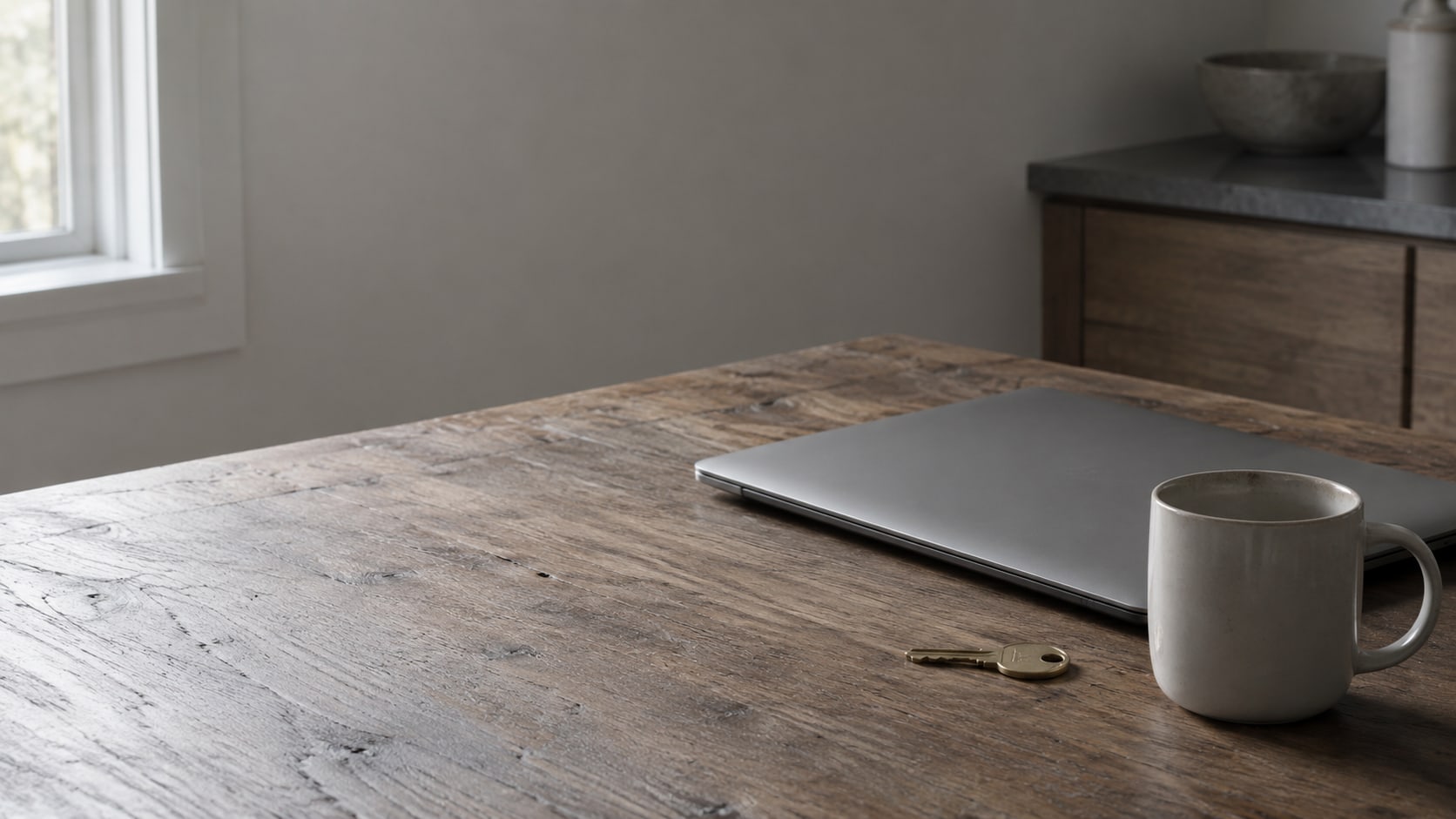 A closed laptop and a small brass house key on a wooden kitchen table beside an empty coffee mug.