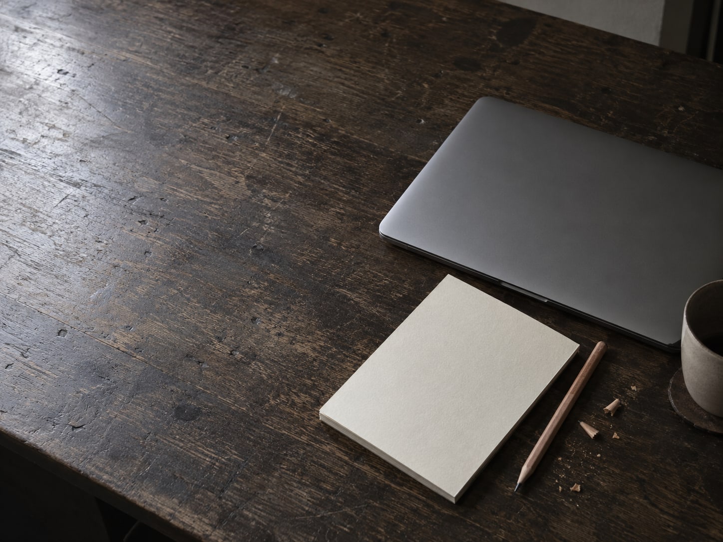 A closed laptop beside a blank notepad and a sharpened pencil on a worn wooden desk.