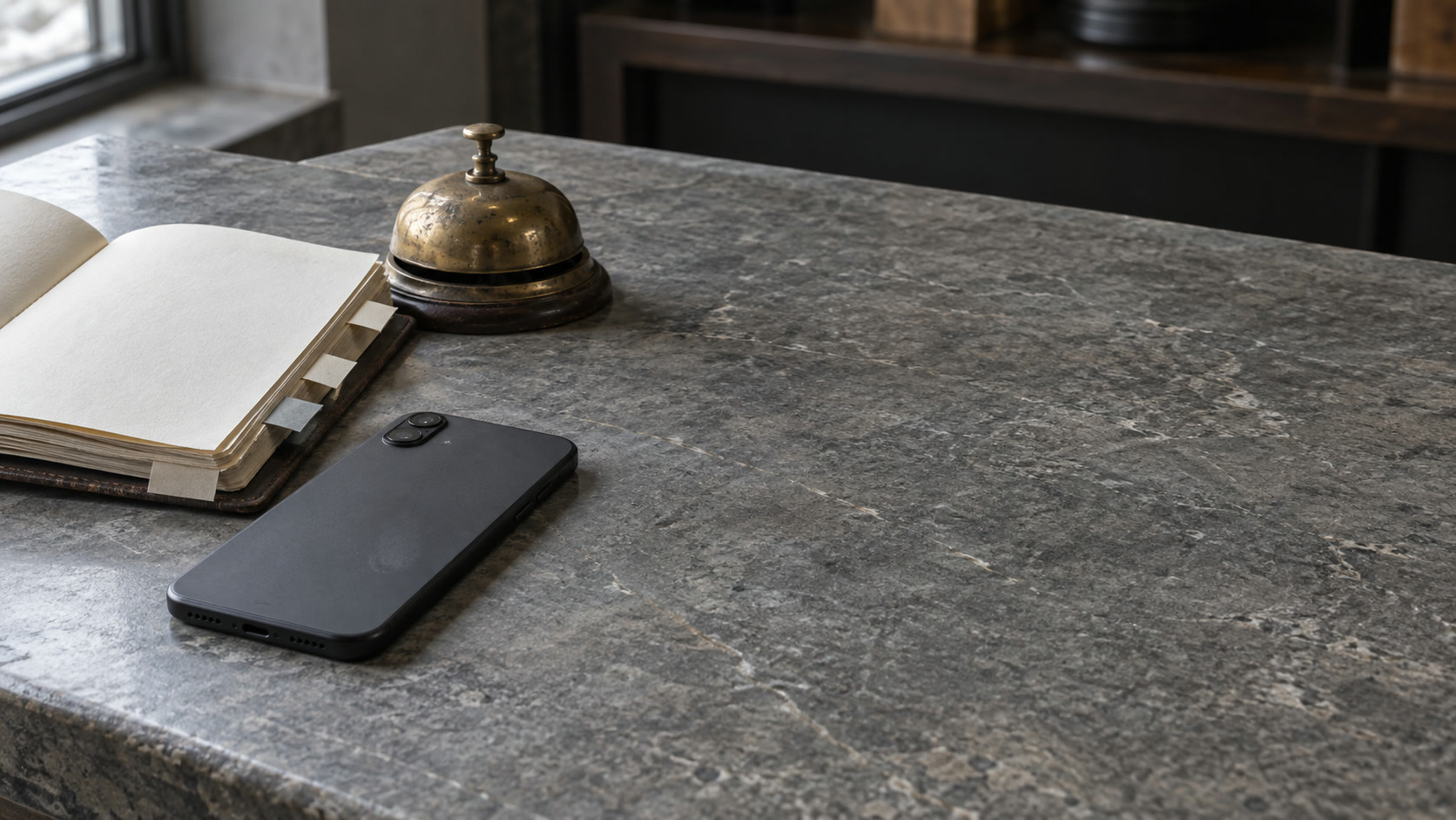 Un teléfono boca abajo sobre un mostrador de piedra junto a una agenda de citas abierta y un timbre de latón.
