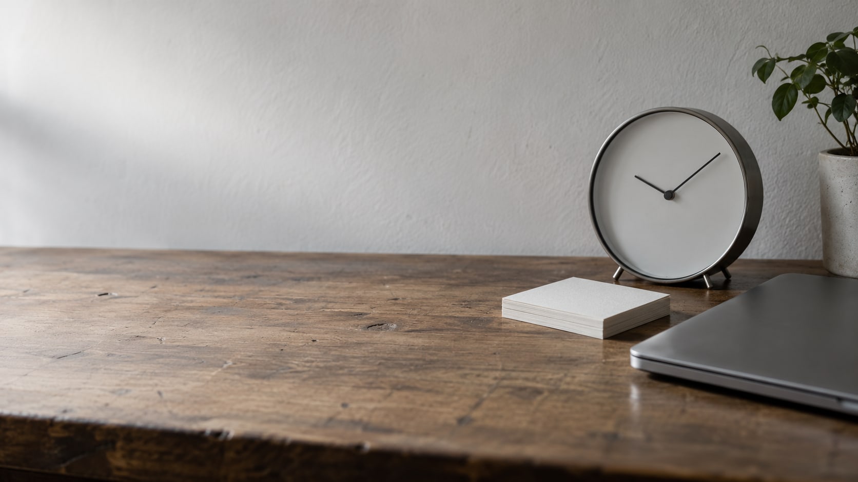 A plain wall clock beside a small stack of blank index cards and a closed laptop on a worn wooden desk.