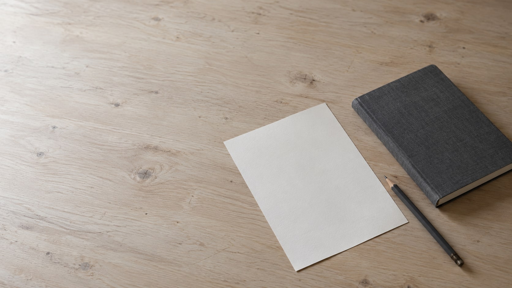 A blank sheet of paper and a pencil lying beside a closed notebook on a pale wooden desk.