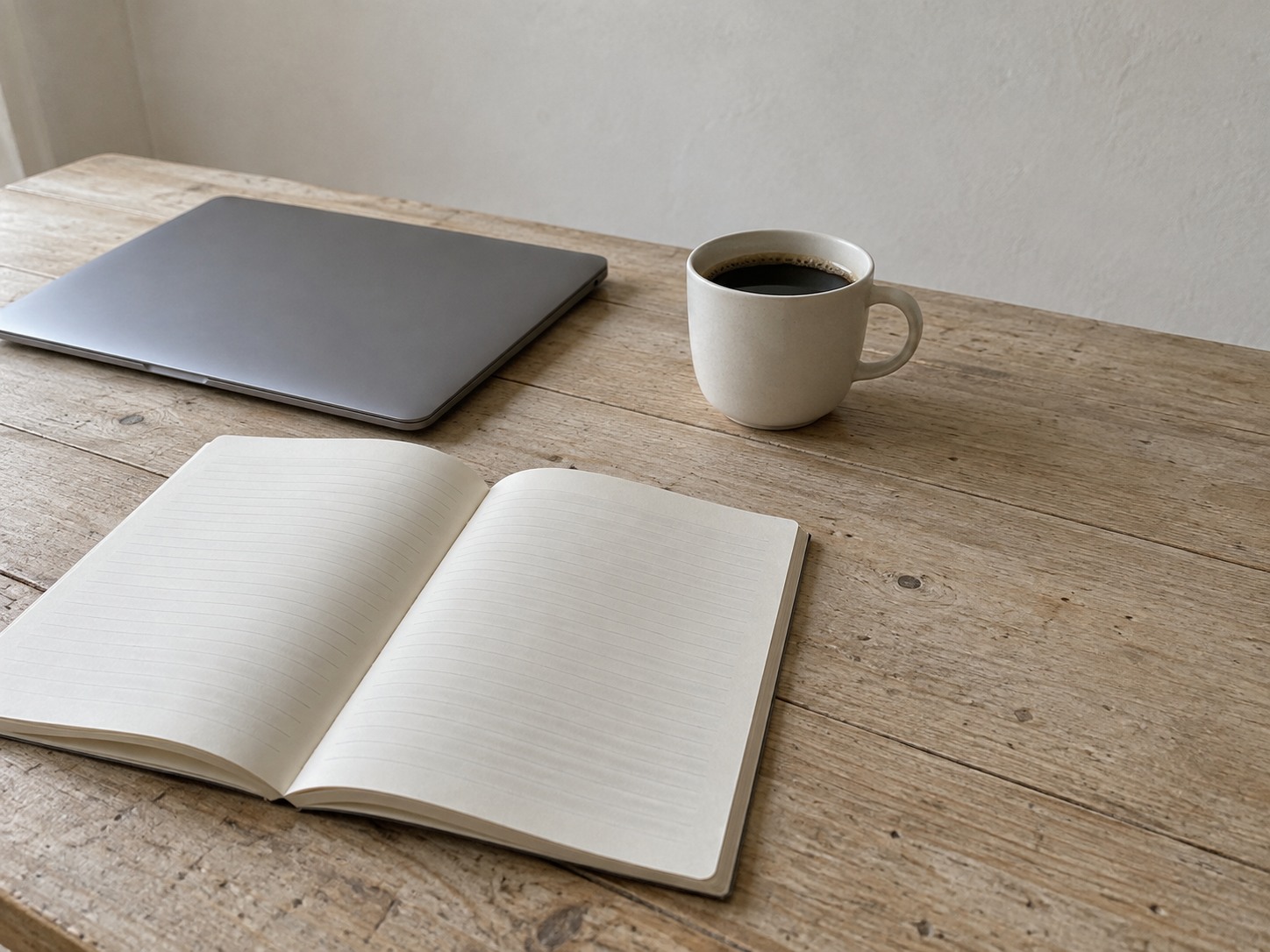 A plain paper notebook open to a page of faint pencil lines, beside a closed laptop and a cup of coffee on a kitchen table.