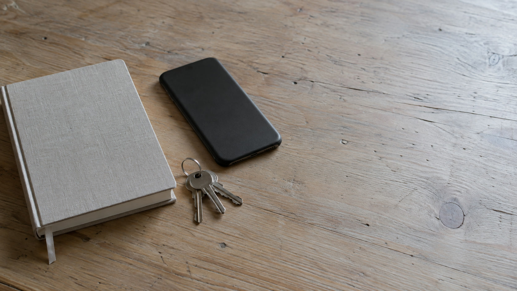 A face-down phone, a small ring of keys and a closed planner arranged on a light wooden desk.