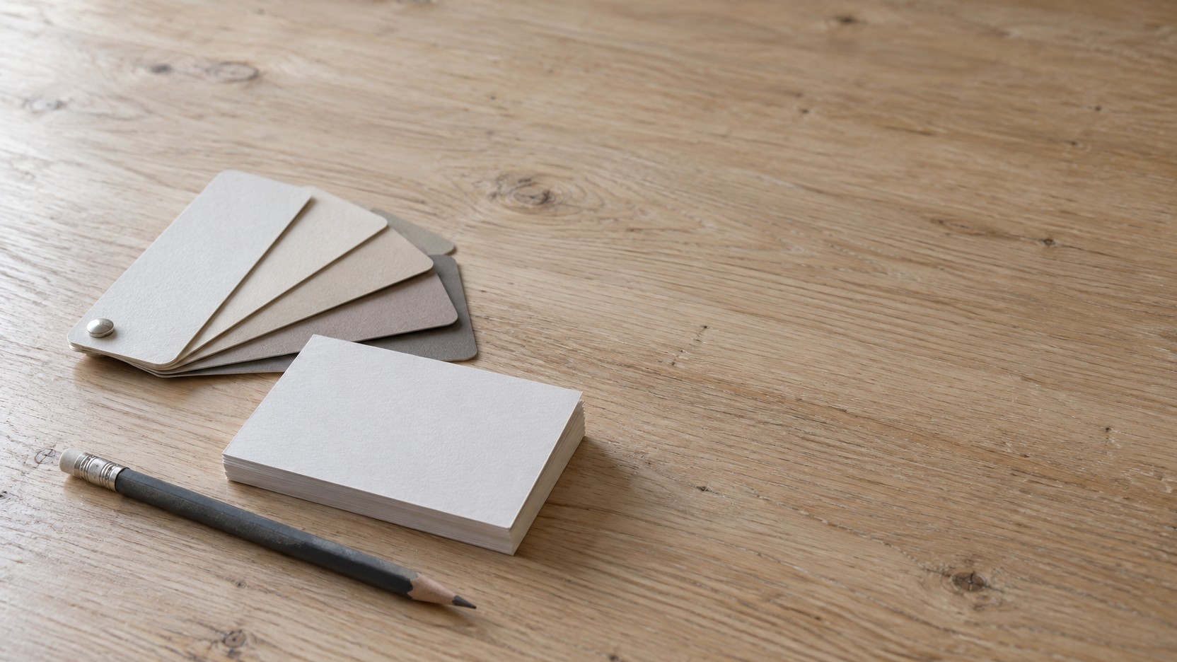 Tarjetas de presentación en blanco, un pequeño abanico de muestras de pintura sin etiquetas y un lápiz sobre un escritorio de madera clara.
