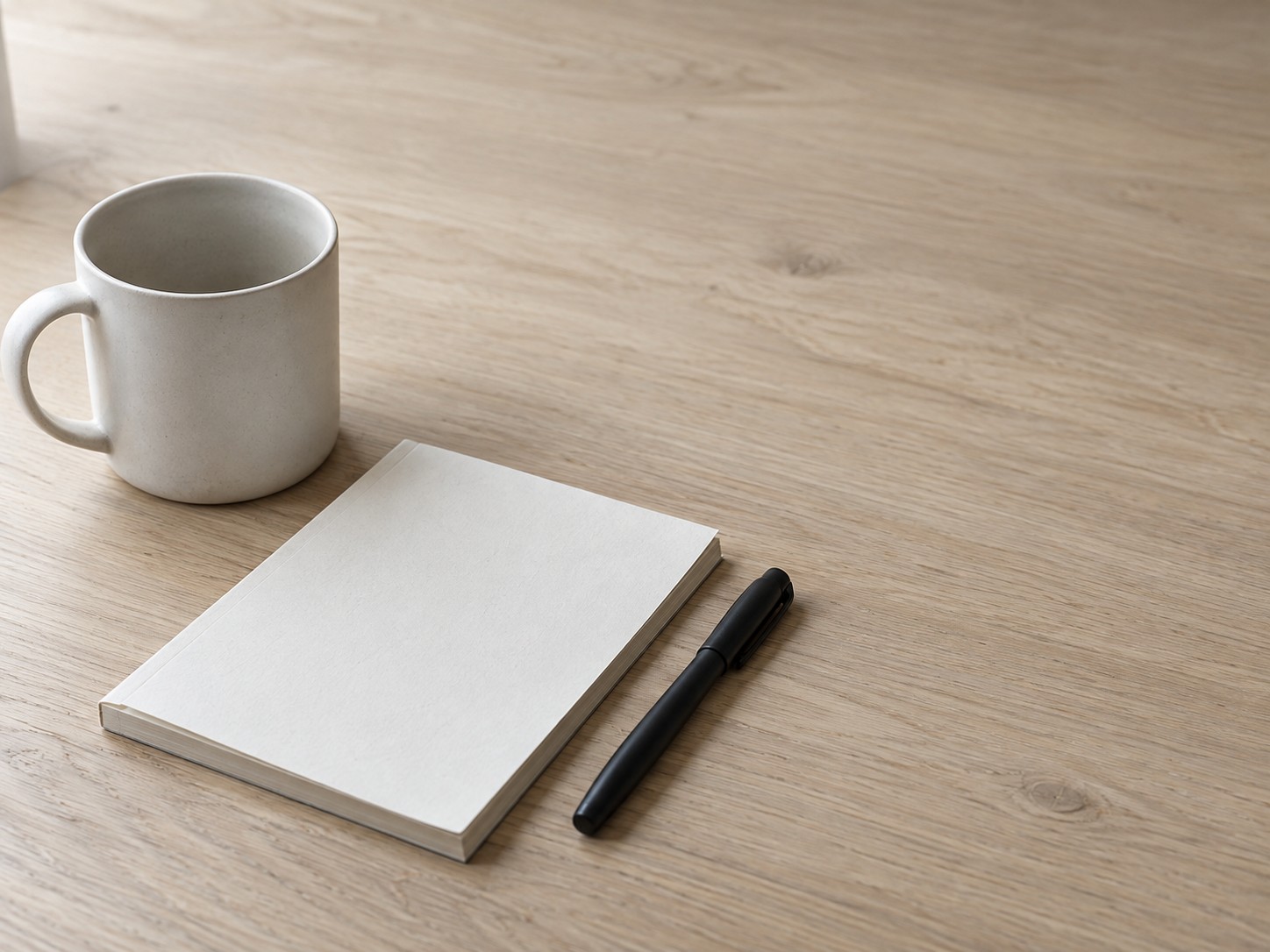 Una sola taza de cerámica, un bloc de notas en blanco y una pluma cerrada sobre un escritorio limpio, sin nada más en el encuadre.