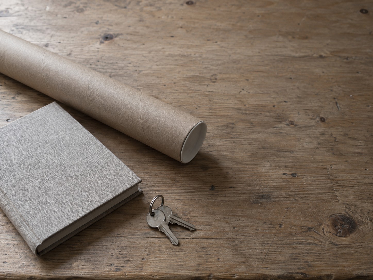 Un tubo de letrero de papel enrollado, un cuaderno cerrado y un pequeño juego de llaves sobre una mesa de trabajo sencilla bajo luz suave de ventana.