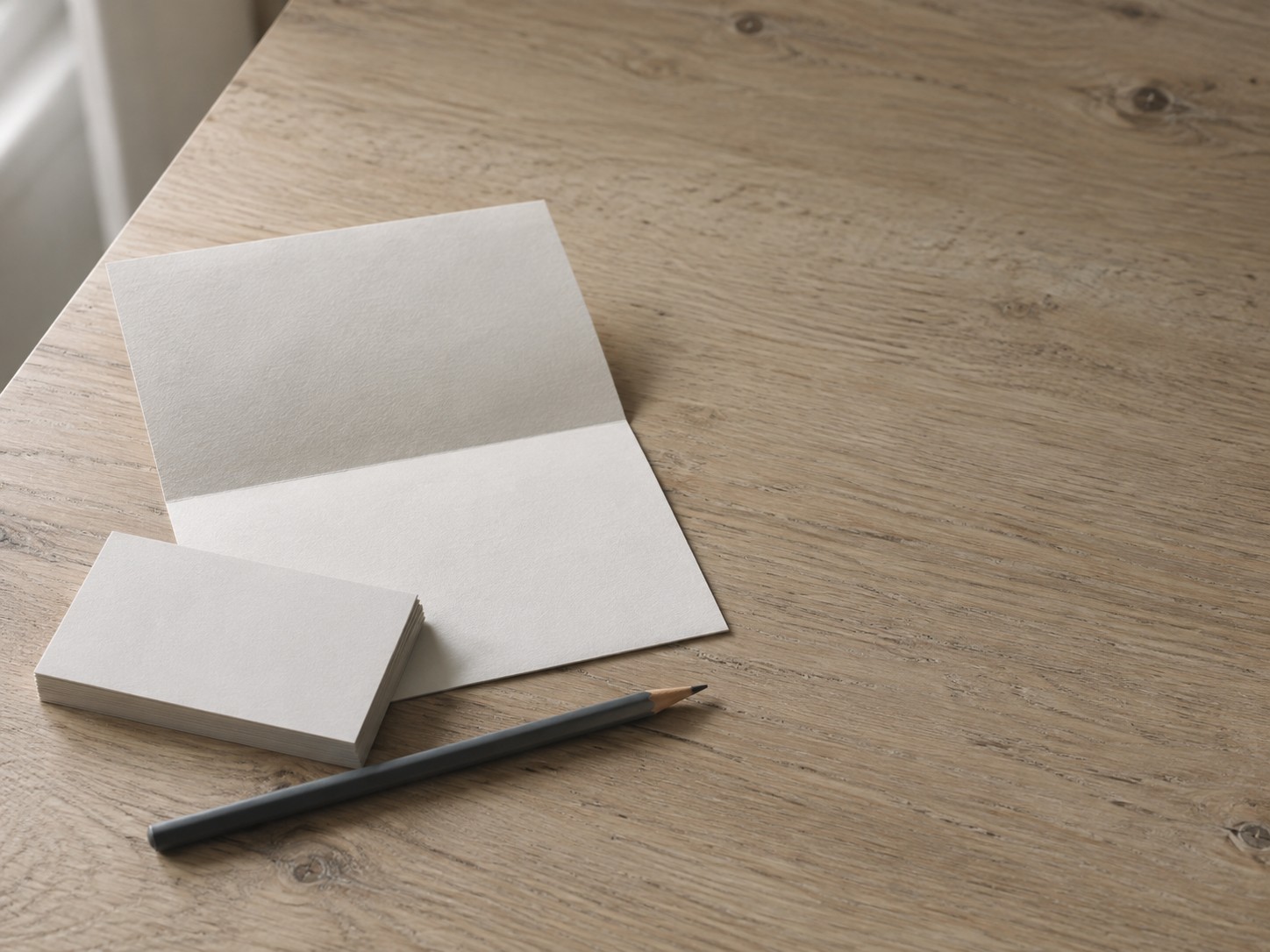 Una pequeña pila de tarjetas de presentación en blanco, un lápiz y una hoja doblada en blanco acomodados con cuidado sobre una mesa de roble clara.