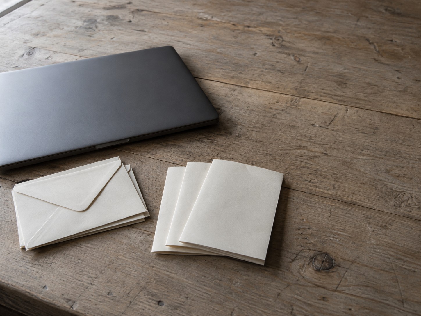Una laptop cerrada junto a una pequeña pila de sobres en blanco y tres cotizaciones dobladas en papel sobre un escritorio de madera gastado.