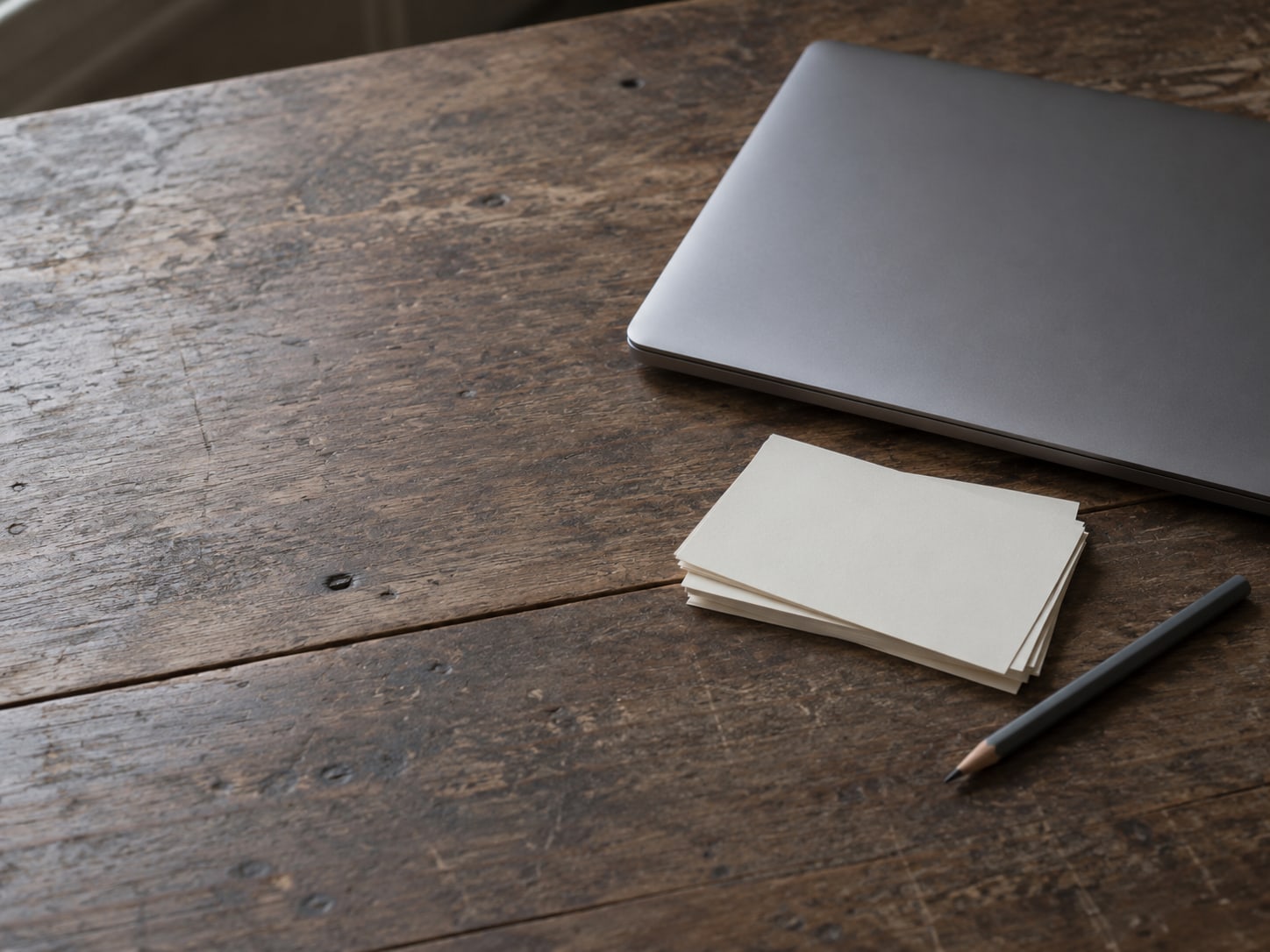 A closed laptop beside a small stack of blank index cards and a pencil on a worn wooden desk.