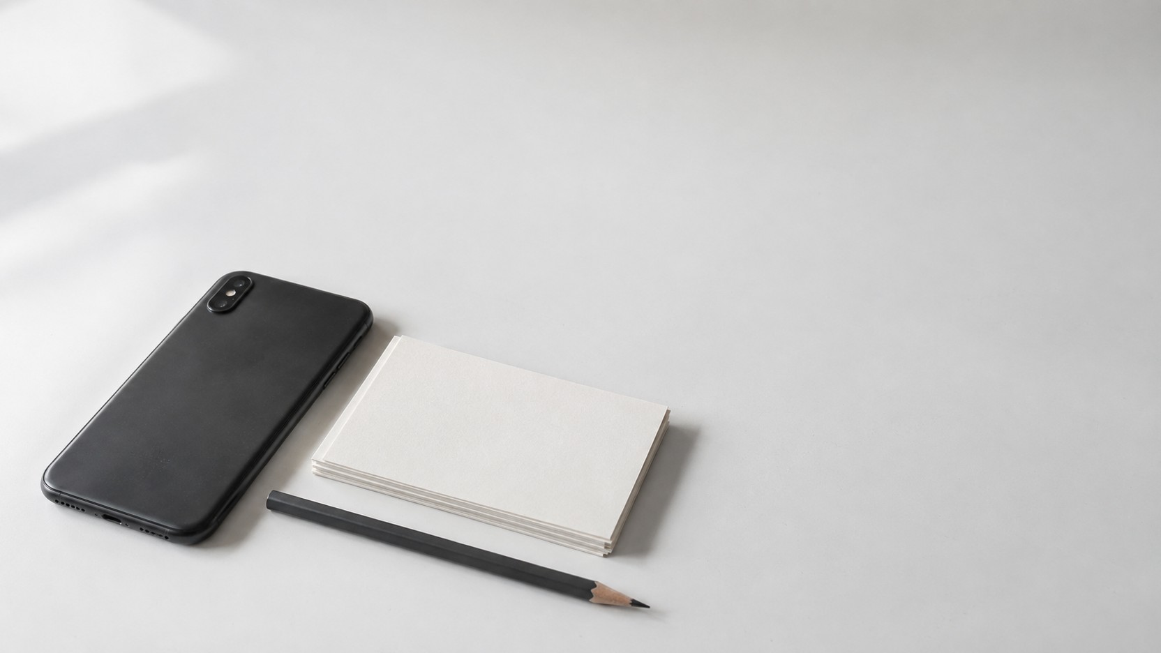 Un smartphone boca abajo junto a un conjunto de tarjetas de wireframe en papel en blanco y un lápiz sobre un escritorio blanco.