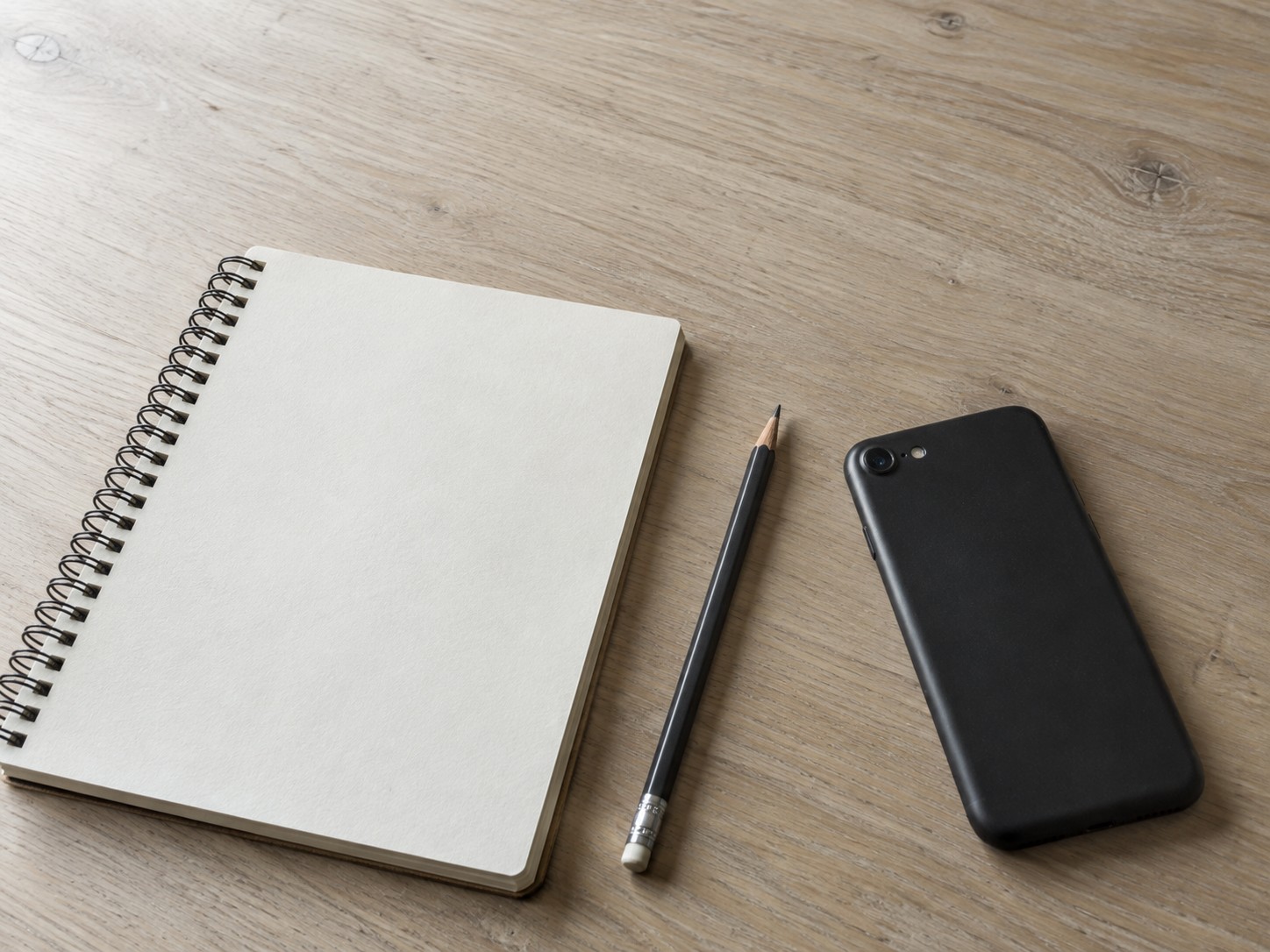 A spiral notebook open to a page of faint pencil marks beside a sharpened pencil and a switched-off phone lying face down.