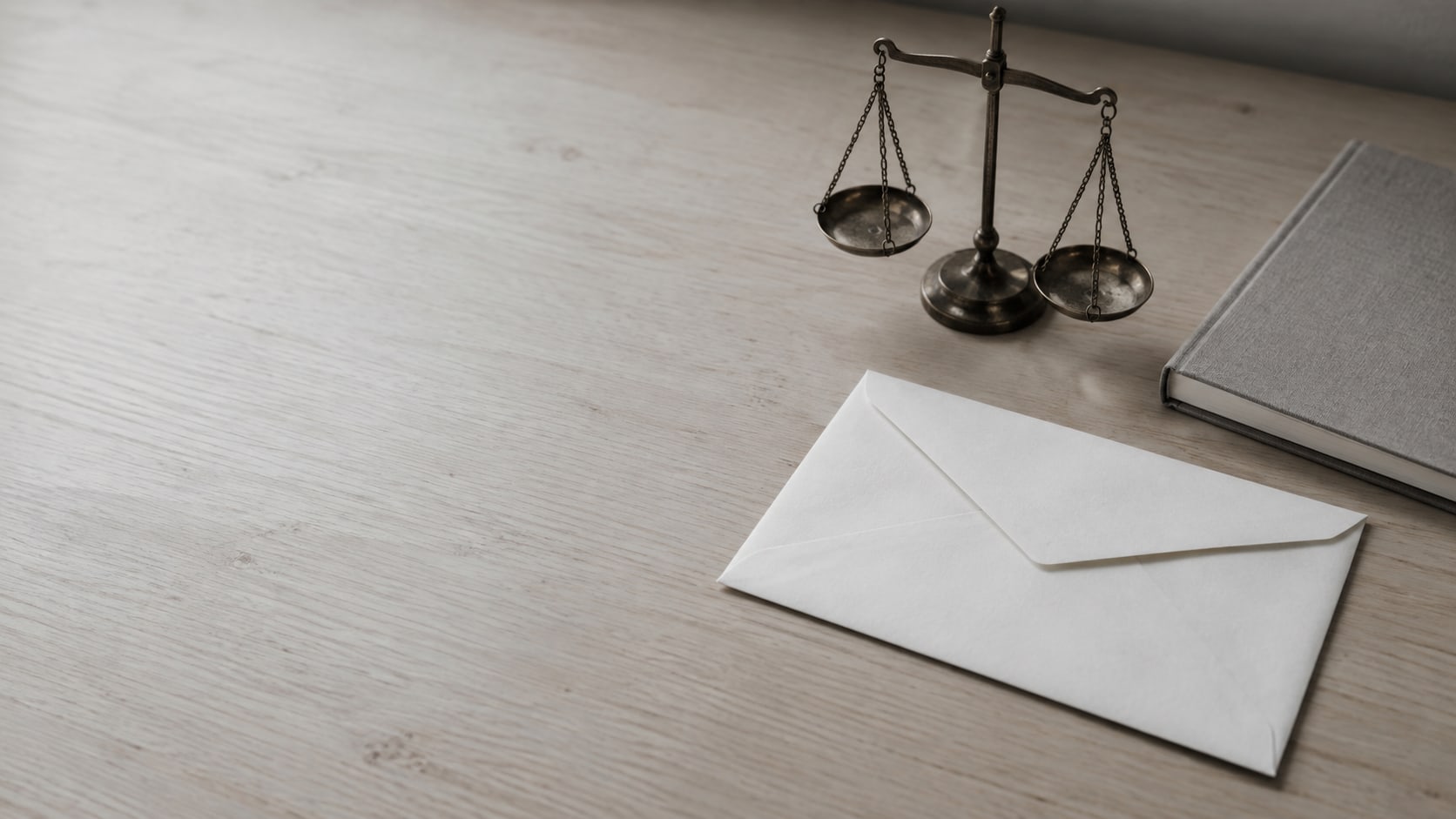 A blank sealed envelope beside a small brass balance scale and a closed notebook on a pale wooden desk.