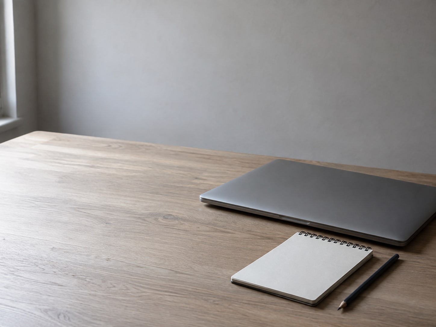 A closed laptop beside a blank notepad and a sharpened pencil on a light oak desk.