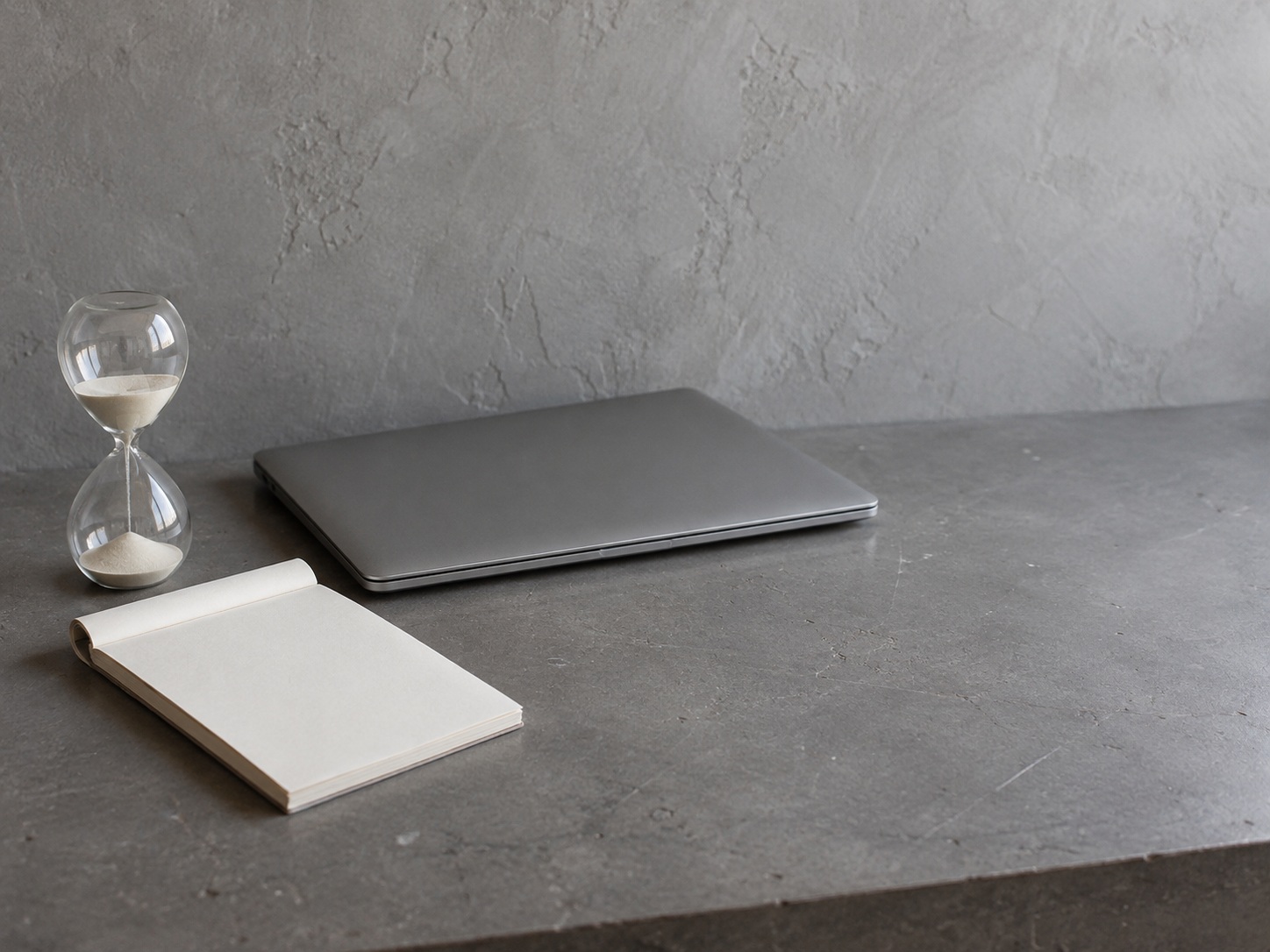 A sand timer beside a folded blank notepad and a closed laptop on a gray desk.