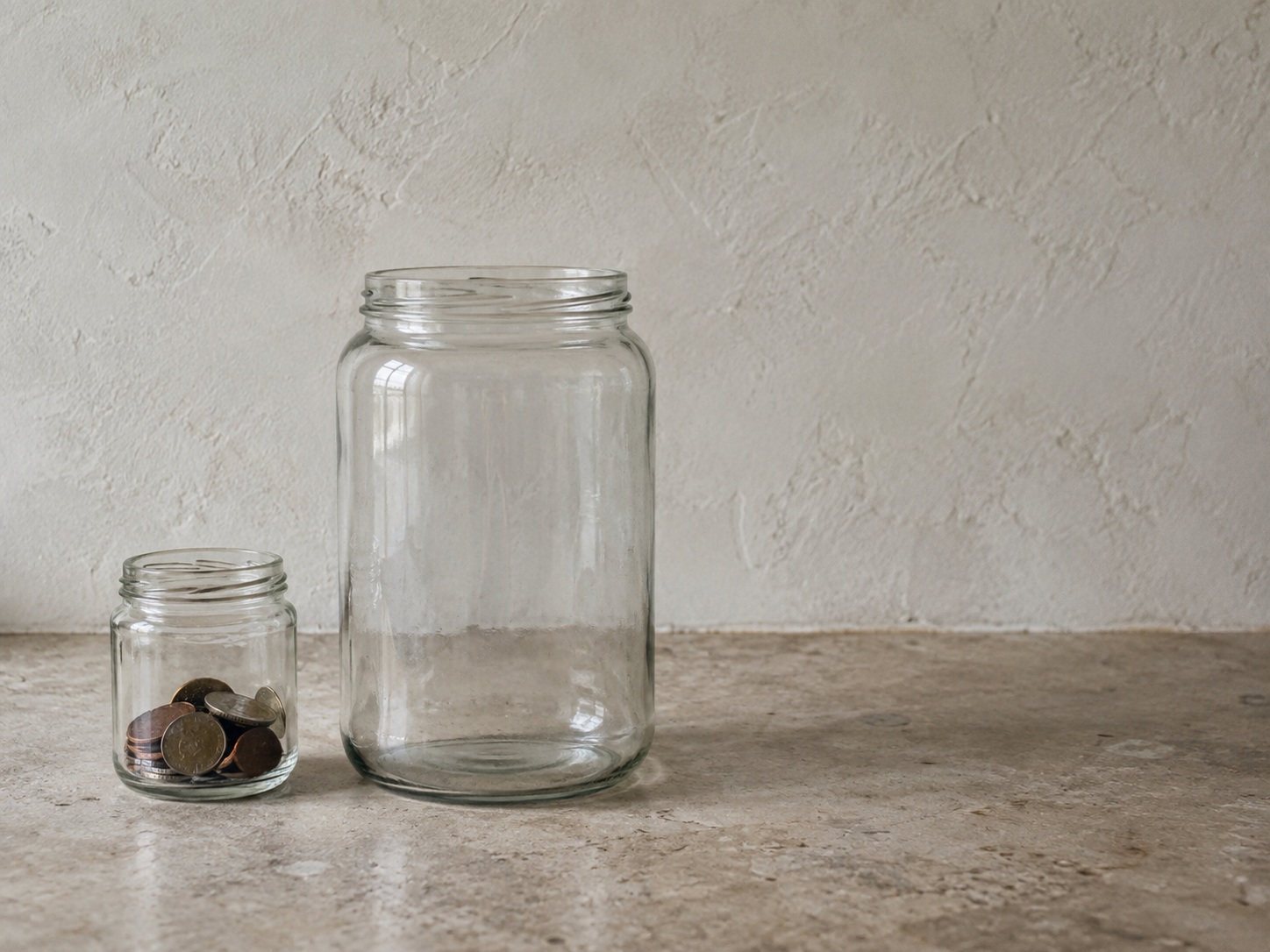 A small glass jar holding a few coins beside a much larger empty glass jar on a kitchen counter.
