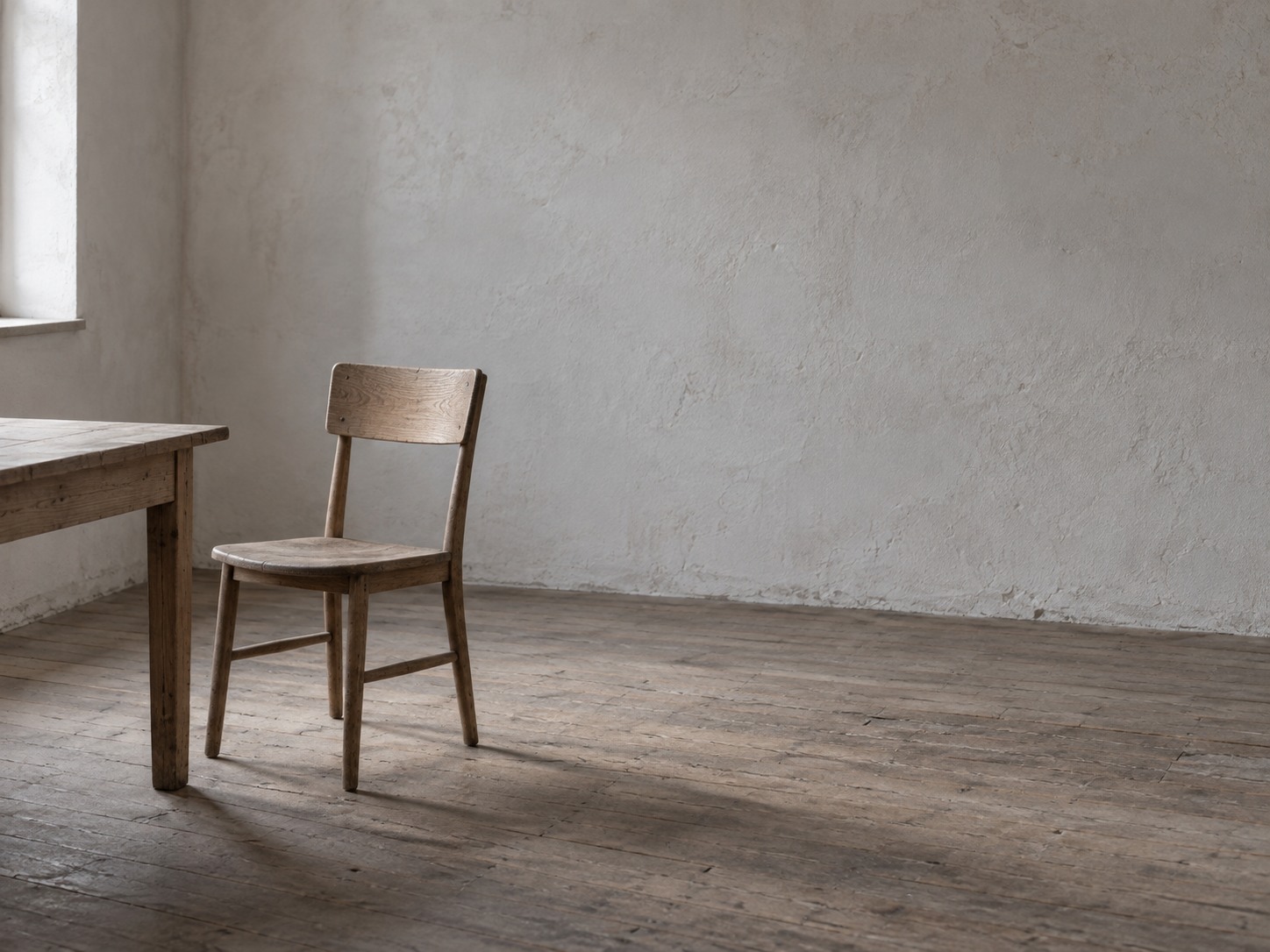 A single empty chair pulled out from a small wooden table in a quiet room with bare walls.